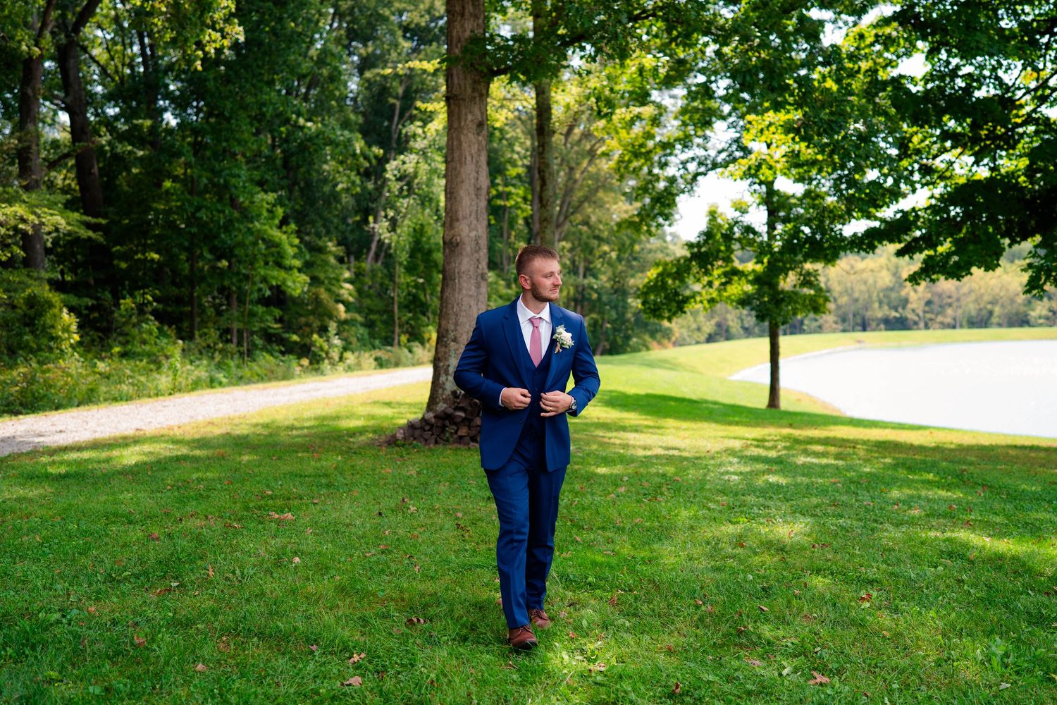 Dapper groom in navy blue suit walks across green lawn at outdoor wedding venue.