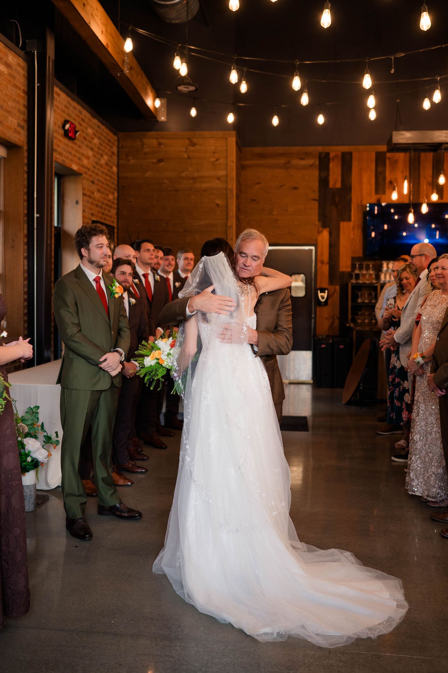 Bride and father share emotional first dance hug at rustic wedding venue with Edison bulb string lights.