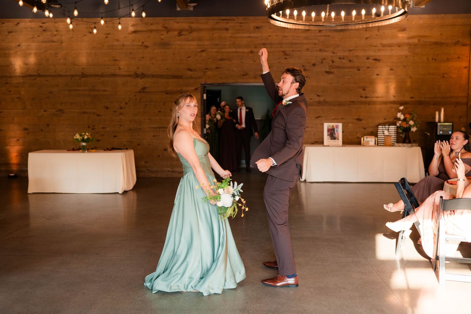 Bridesmaid in teal gown dances with groomsman in brown suit at rustic wedding reception venue.
