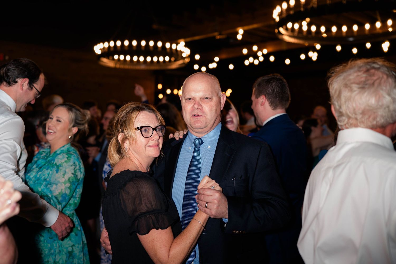 Couple dances together at lively wedding reception under warm chandeliers surrounded by guests.