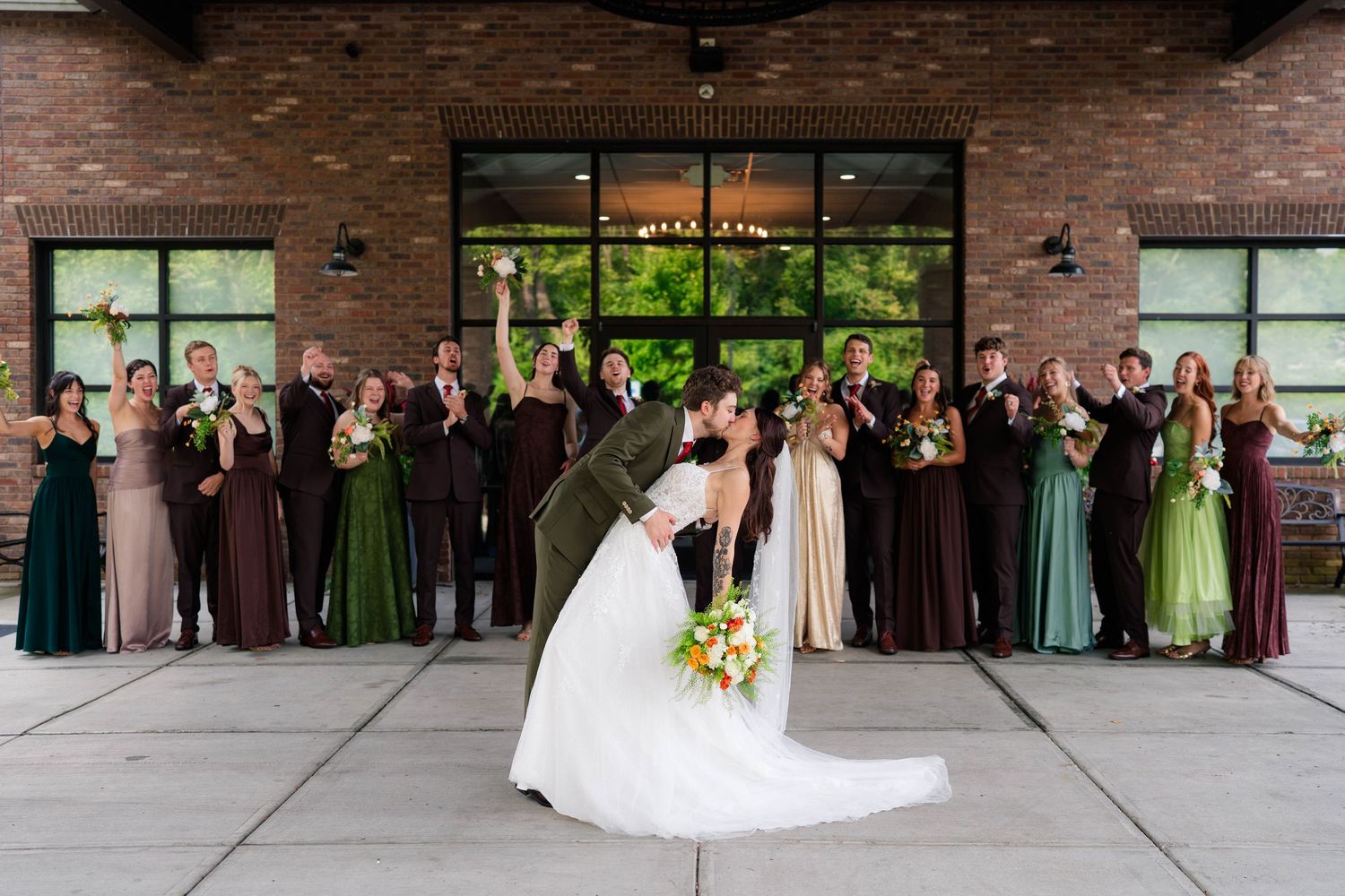 Bride and groom kiss while wedding party in green and brown attire celebrates behind them outdoors.