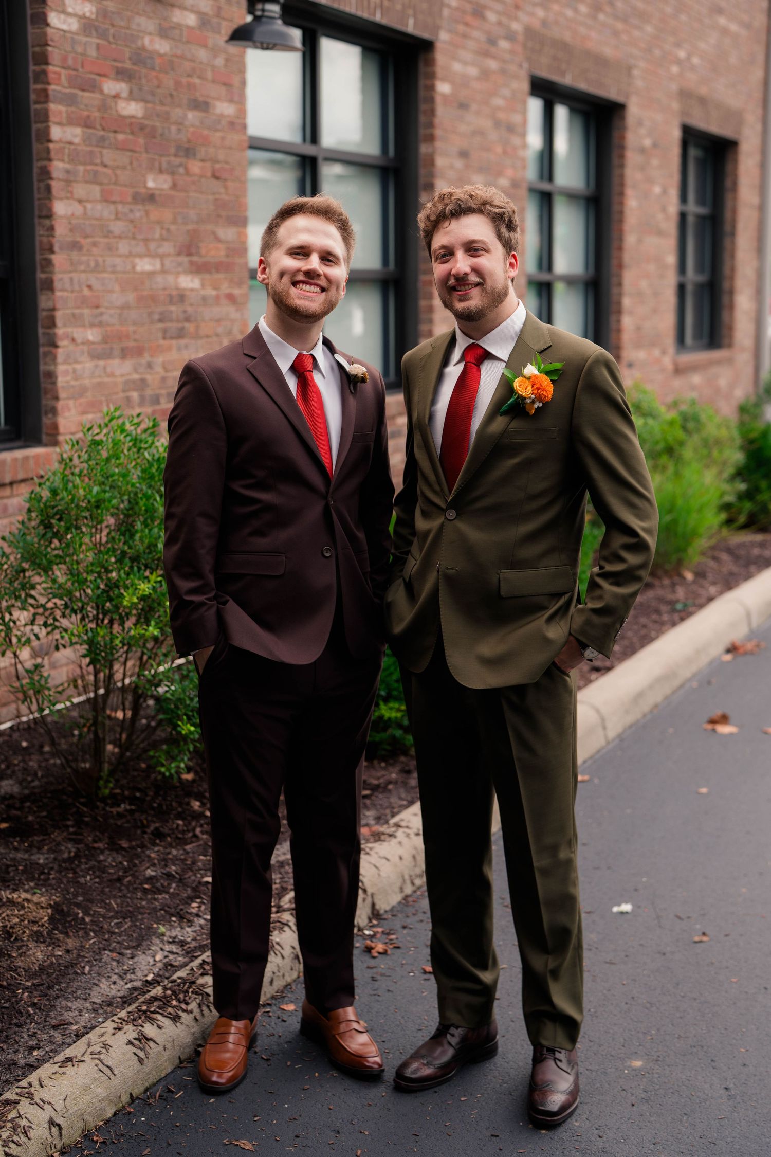 Two groomsmen in olive green and burgundy suits with boutonnieres pose outside brick wedding venue.