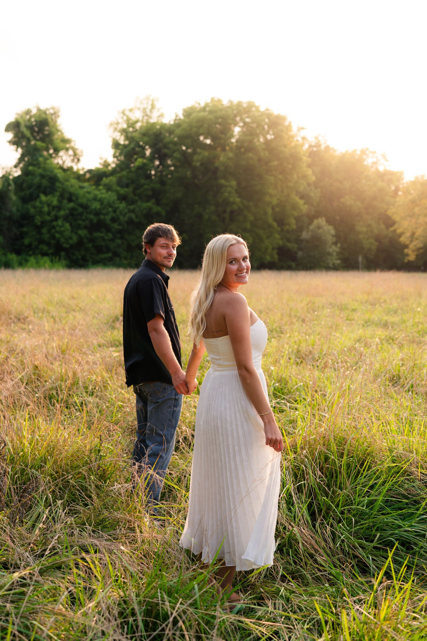 A smiling blonde woman in a white maxi dress holds hands with a man in a field during golden hour sunset.