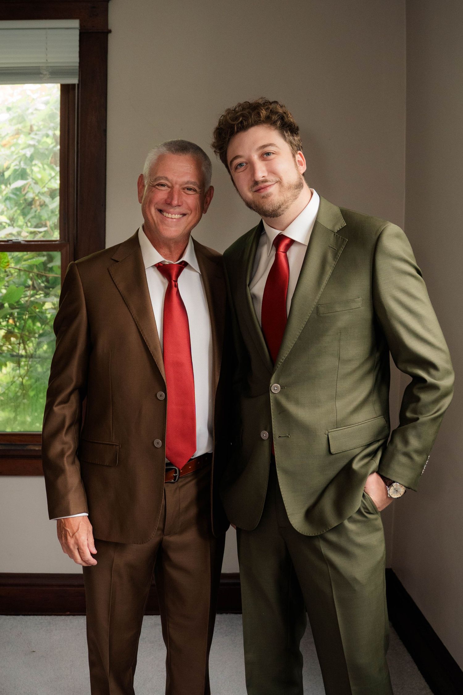 Two men in suits with red ties pose together smiling near a window.