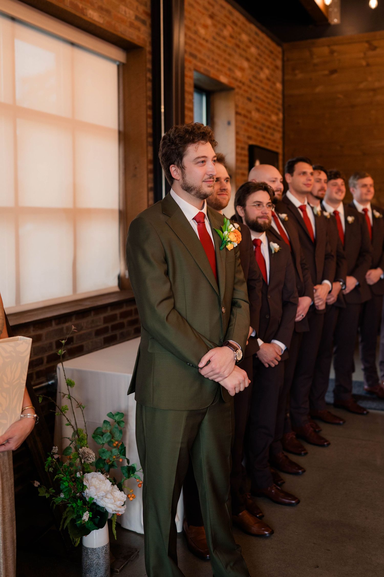 Groom in green suit smiles at altar flanked by groomsmen in dark suits with red ties.