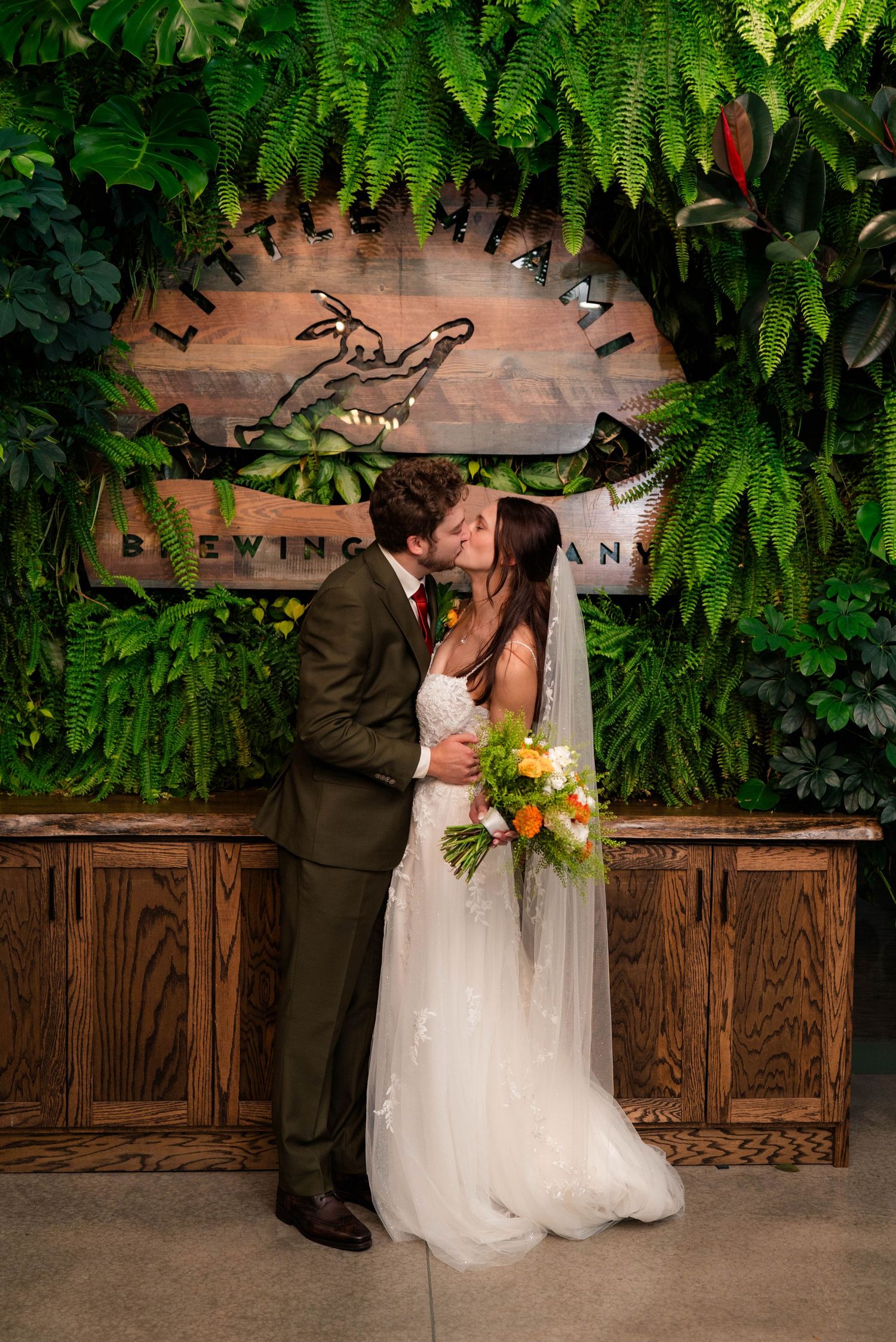 Bride and groom kiss in front of a lush green plant wall at Little Miami Brewing Company.