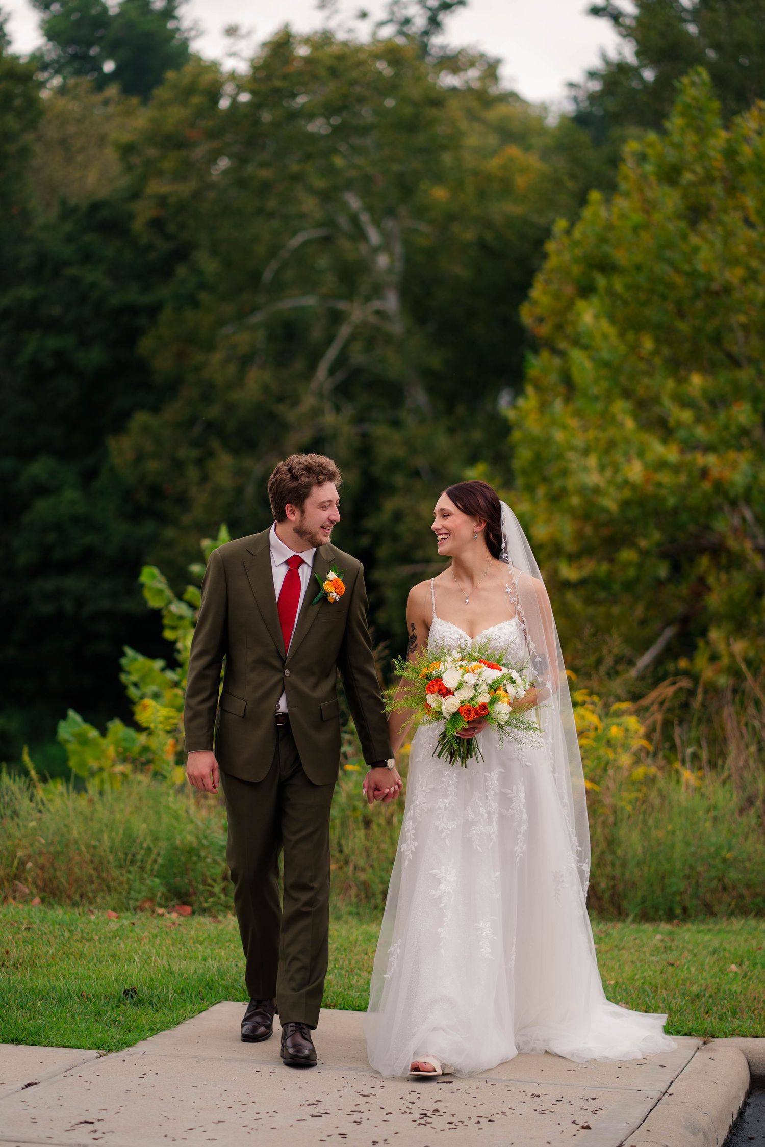 Bride and groom walk hand-in-hand on outdoor path surrounded by lush autumn foliage at fall wedding.