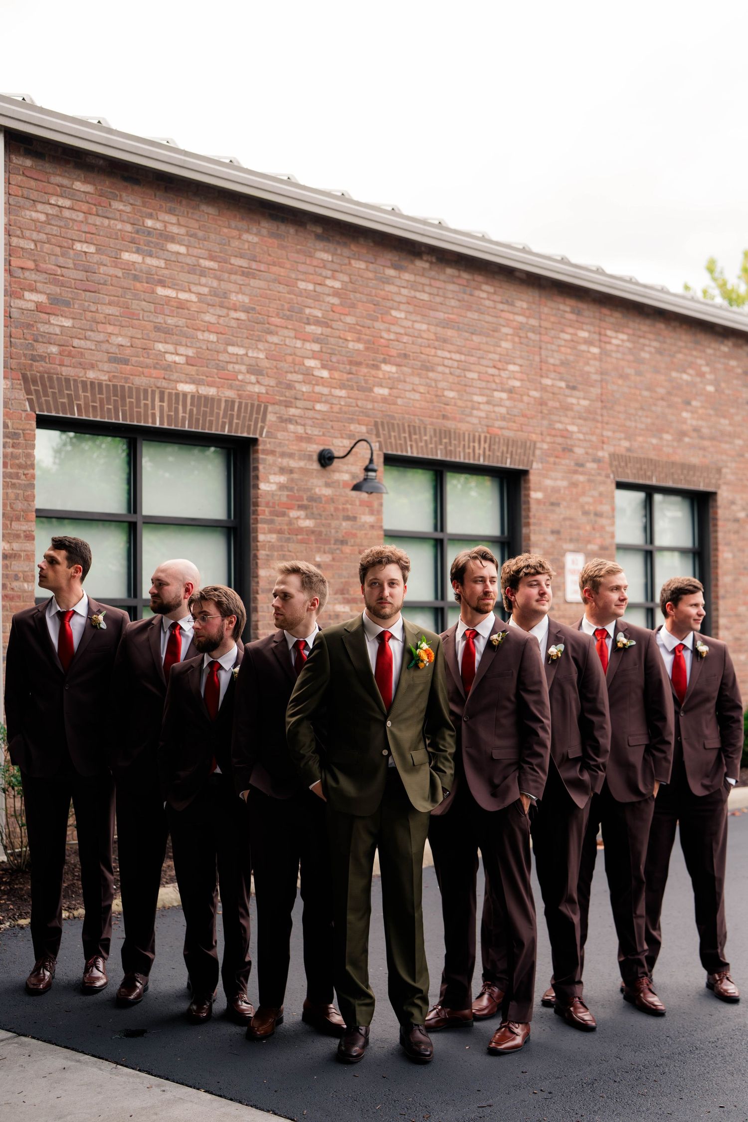 Groom in olive suit stands with groomsmen in dark brown suits and red ties outside a brick building.