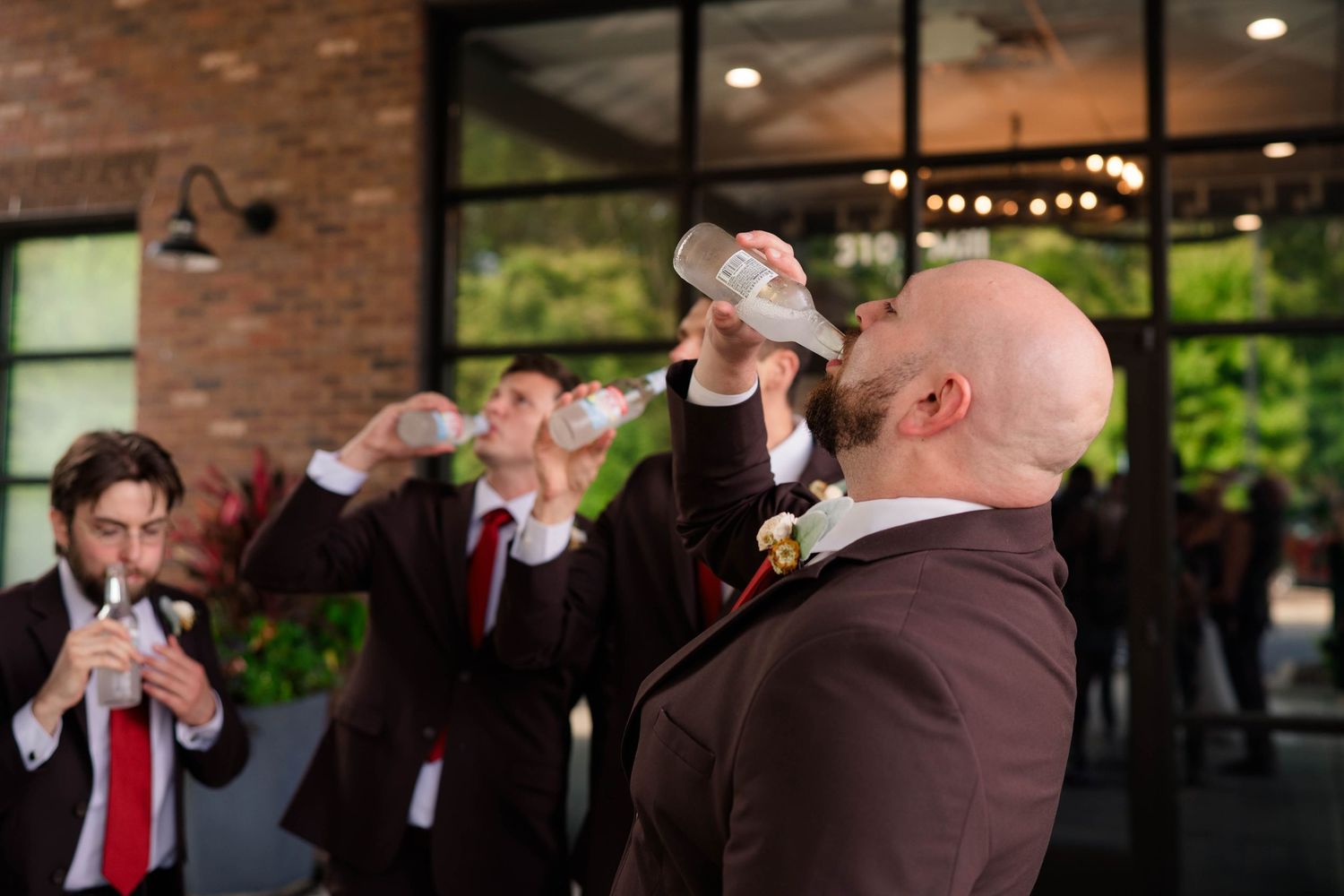 Groomsmen in brown suits with red ties toasting drinks before wedding ceremony.