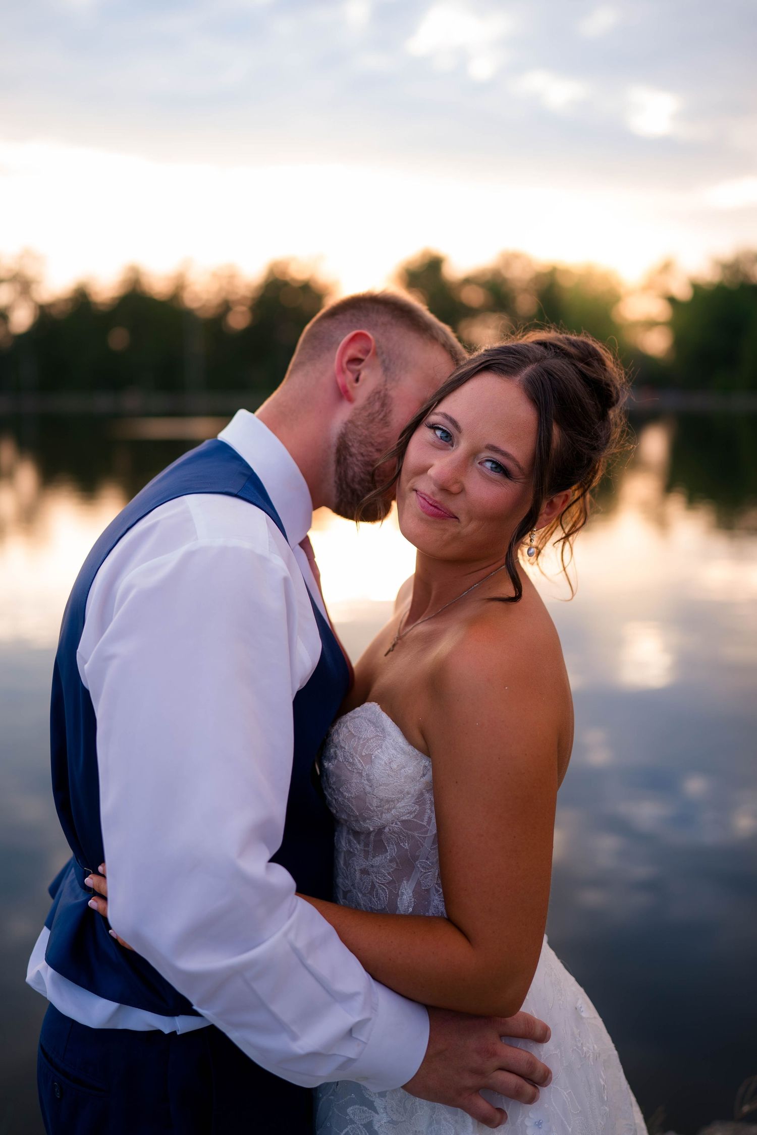 Groom in navy vest kisses bride's cheek as she smiles at camera during golden hour by a serene lakeside.
