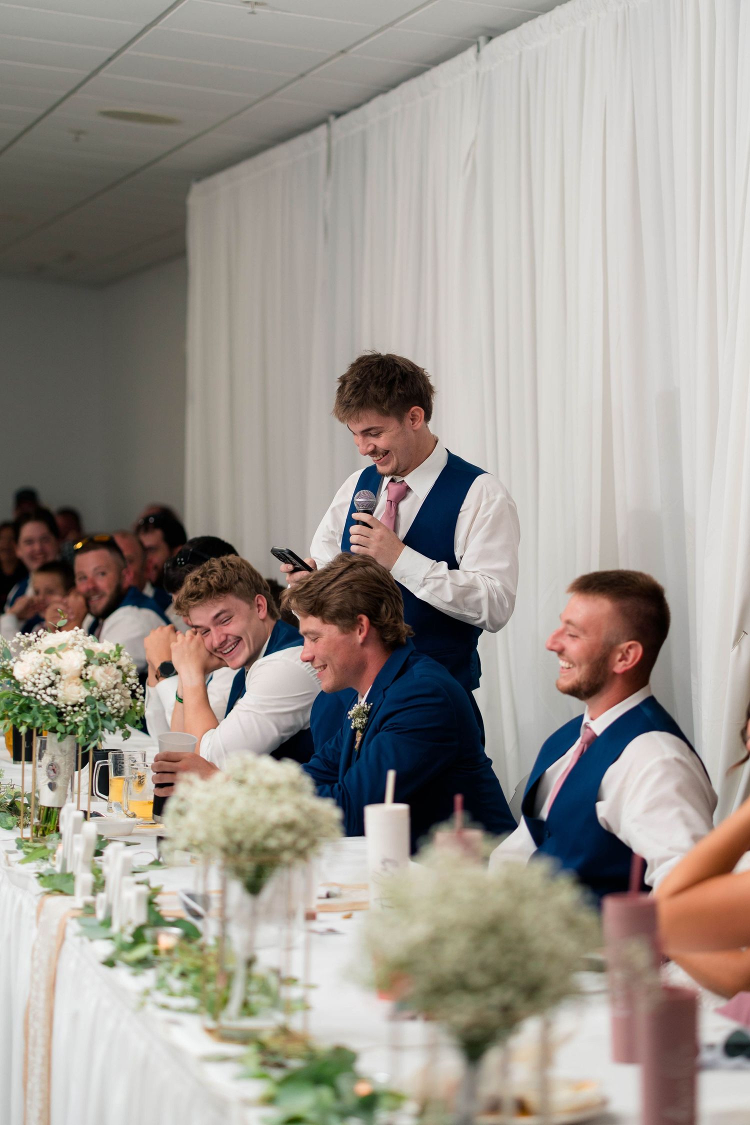 Best man delivers speech at wedding reception as groomsmen laugh at head table with floral decor.
