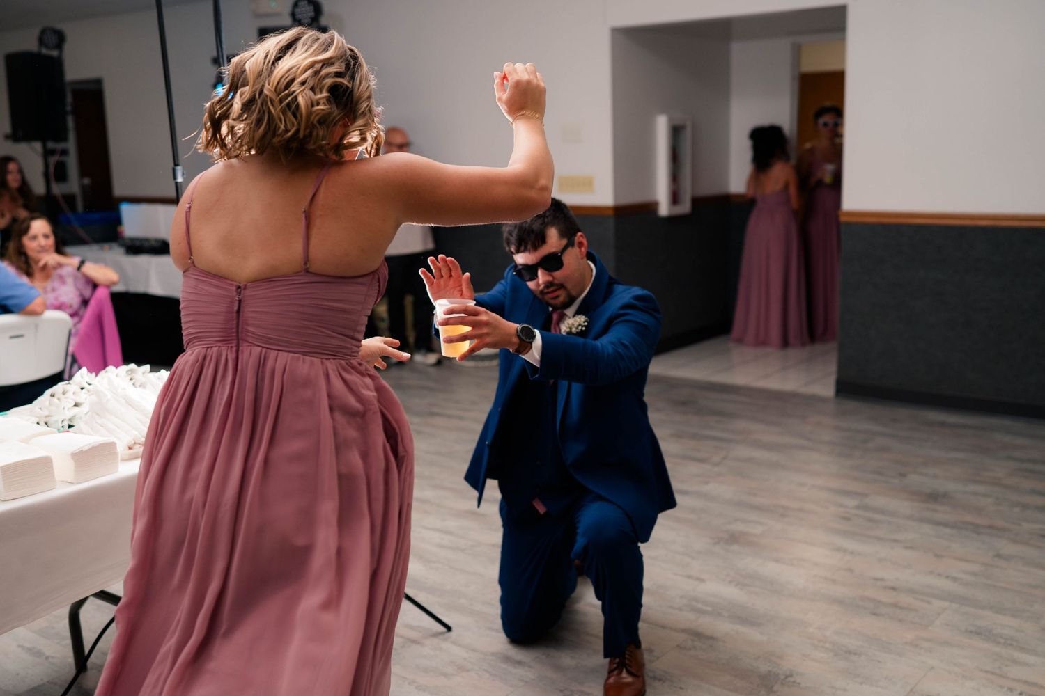 Groomsman in navy suit dancing with bridesmaid in pink dress at indoor wedding reception.