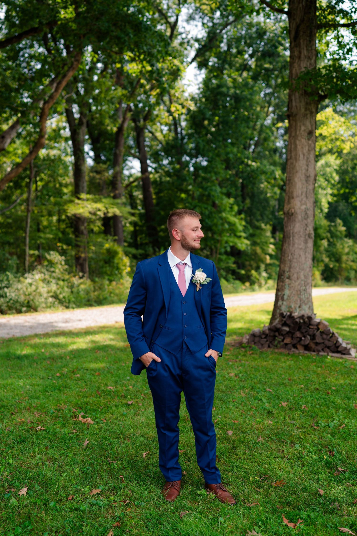 Handsome groom in navy blue three-piece suit with pink tie and boutonniere standing on green lawn.