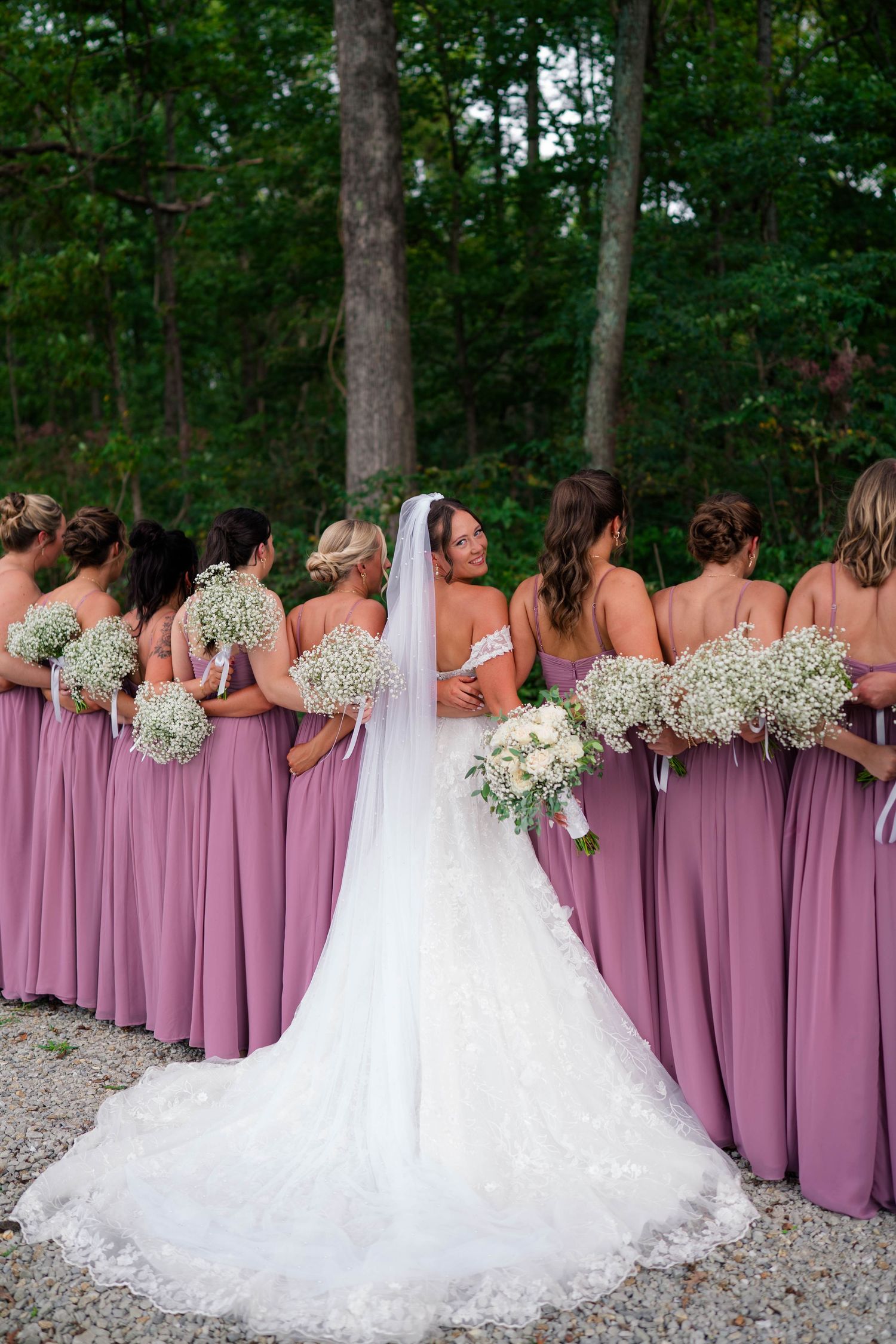 Bride in lace wedding gown with cathedral veil poses from behind with bridesmaids in mauve dresses holding bouquets.