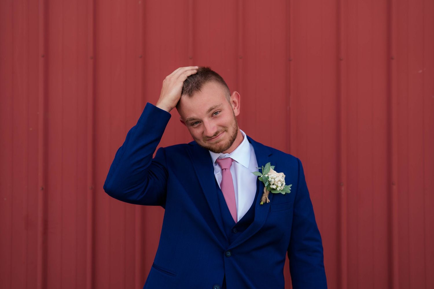 Groom in navy blue suit with pink tie and boutonniere posing against red barn wall.