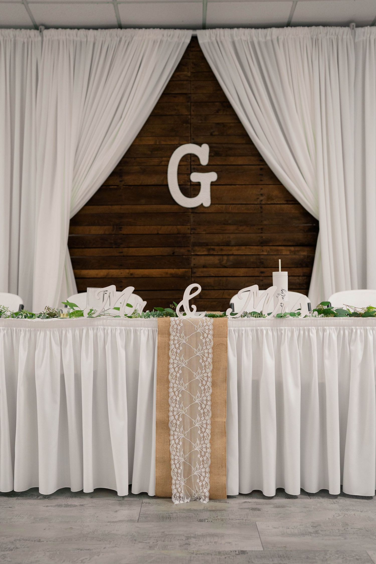 Elegant wedding head table with white draping, rustic wood backdrop, monogram letter G, burlap lace runner, and greenery.