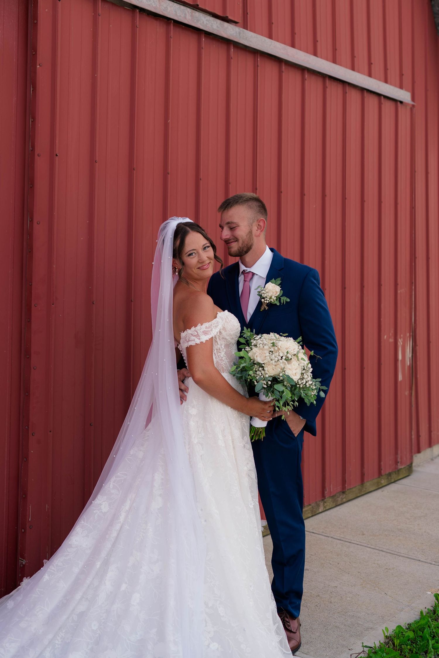 Bride in lace off-shoulder gown and groom in navy suit pose together against a red barn wall.