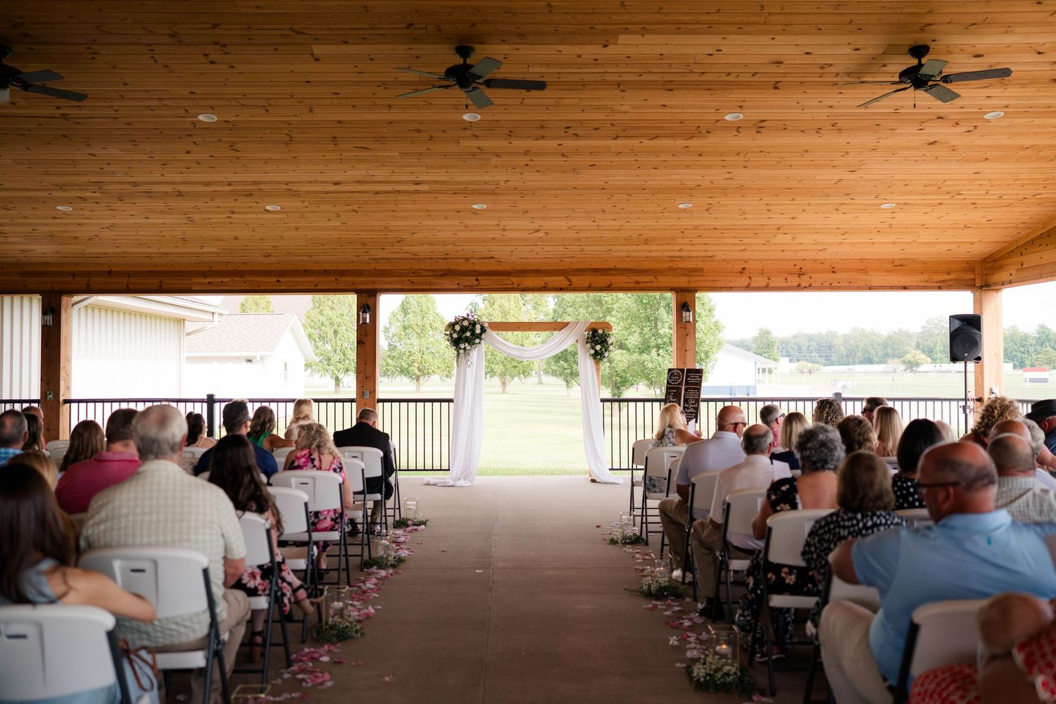 Outdoor wedding ceremony under wooden pavilion with guests seated facing floral arch altar.