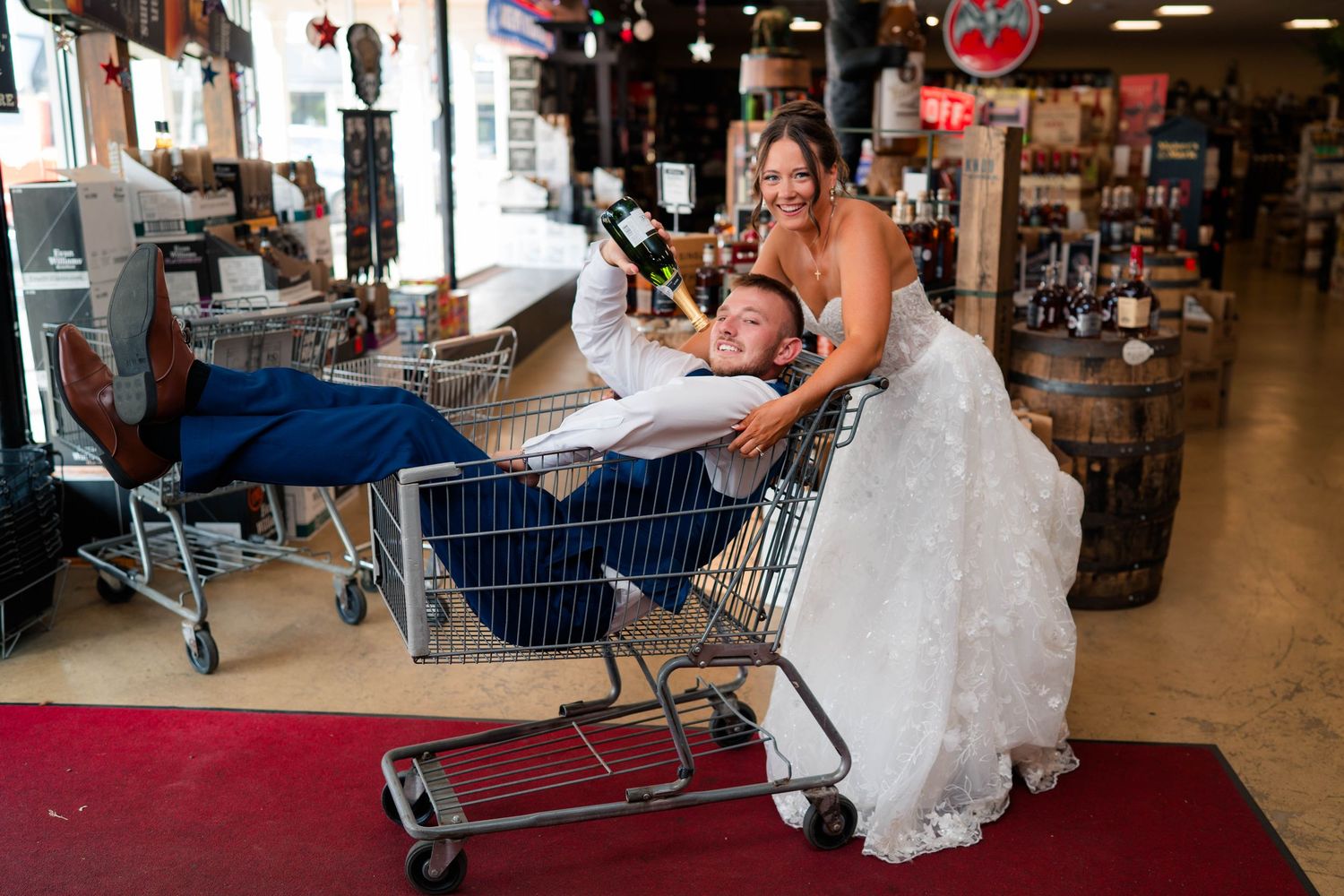 Laughing bride in white gown pushes groom in a shopping cart inside a rustic store.
