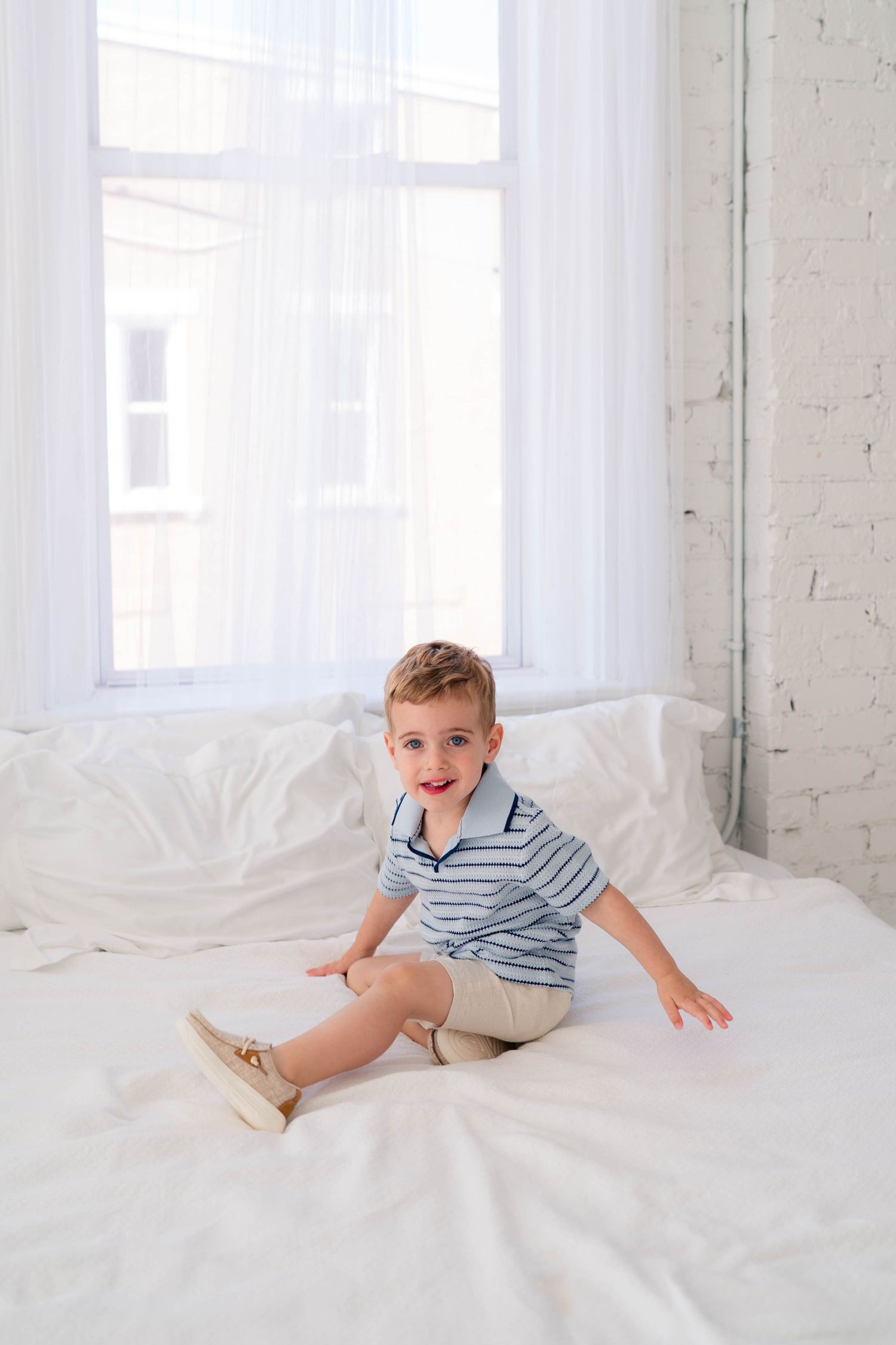 Smiling toddler boy in striped polo shirt sitting on a white bed near bright windows.