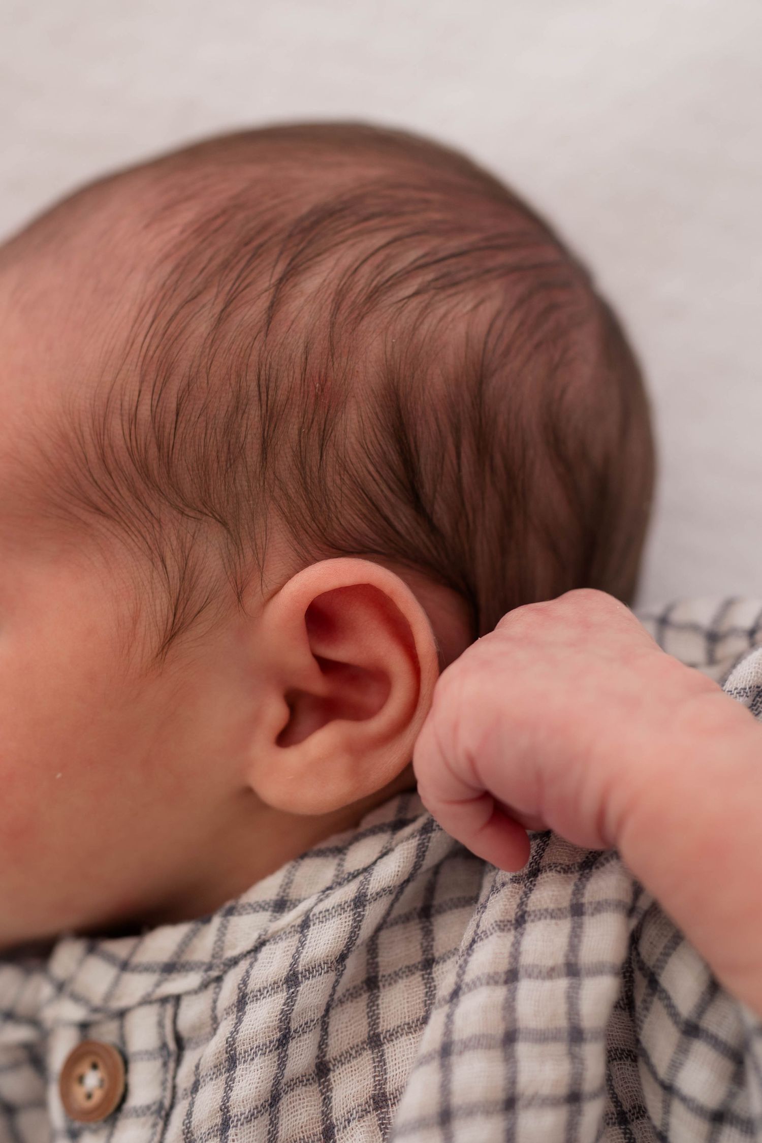 Close-up of a newborn baby's ear and tiny fist, wearing a plaid onesie, lying on a white surface.