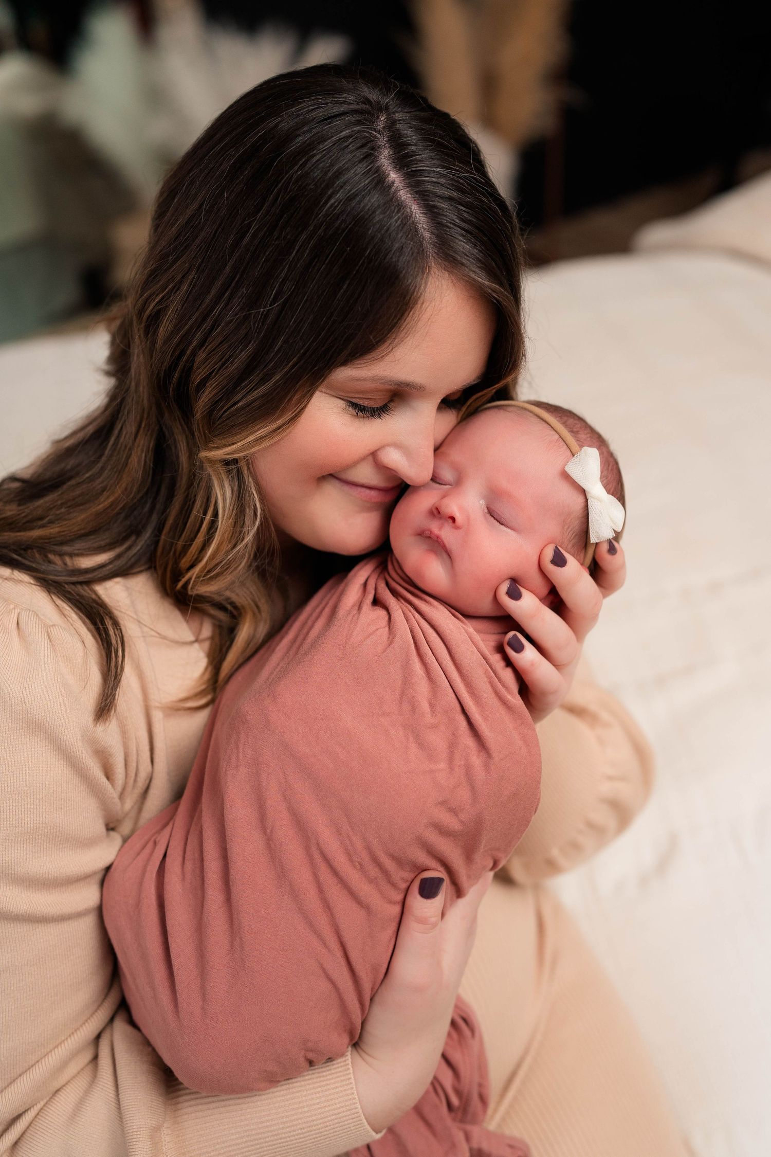 Dark-haired mother smiles cheek-to-cheek with swaddled newborn in dusty rose wrap with bow.