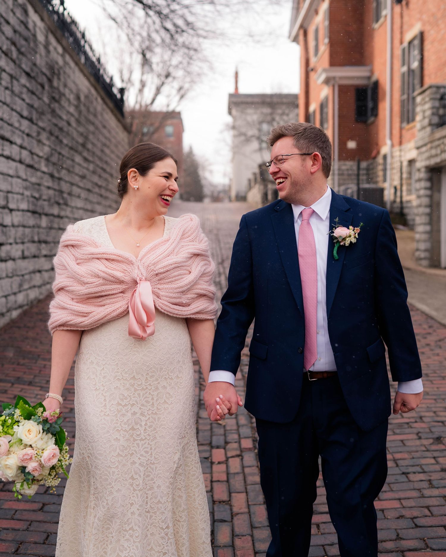 Smiling bride and groom walk hand-in-hand on a cobblestone alley, she holding a bouquet in a pink knit wrap.