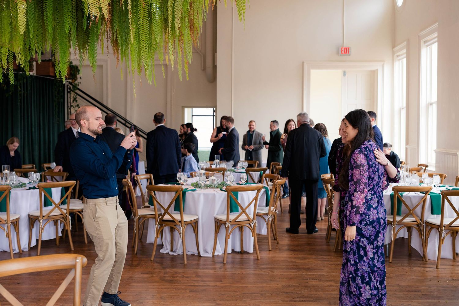 Photographer captures elegant event venue with hanging greenery, round tables, and guests mingling at a formal gathering.