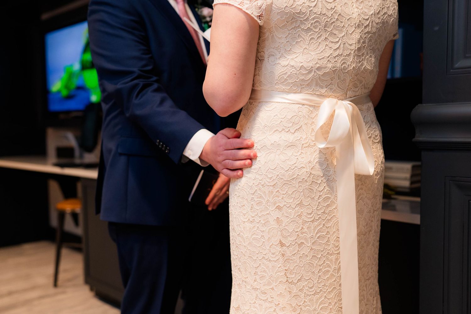 Close-up of groom gently holding brides waist in lace wedding gown with satin bow detail.
