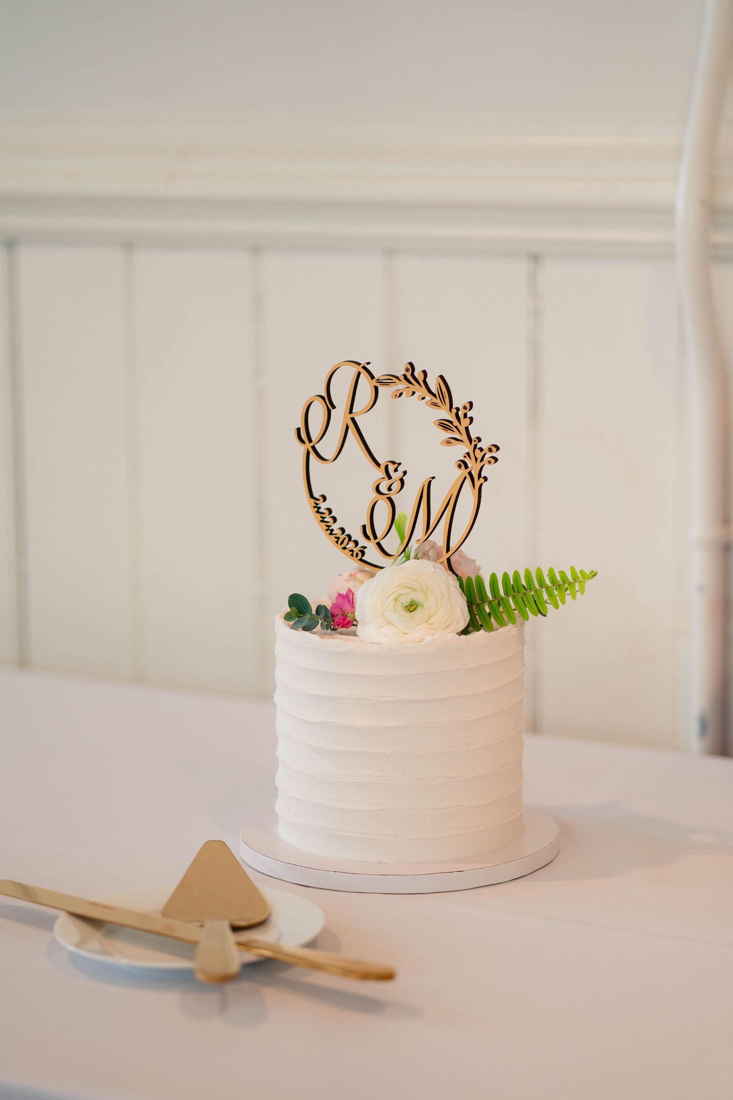 Elegant white single-tier wedding cake with wooden R&M monogram topper and fresh flowers on white table.