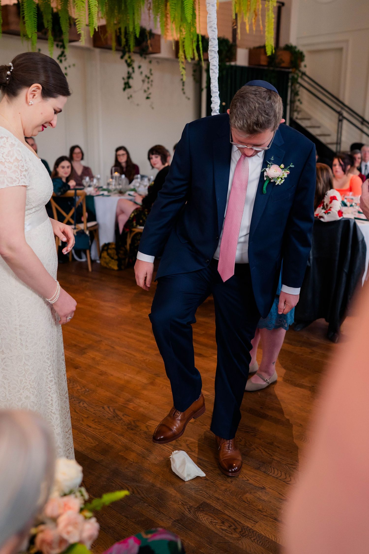 Groom in navy suit stomps on glass during traditional Jewish wedding ceremony while bride watches smiling.