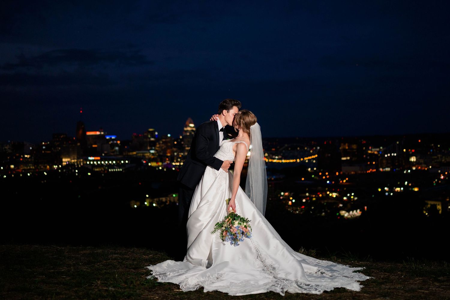 Bride and groom kiss romantically against a stunning city skyline at night.