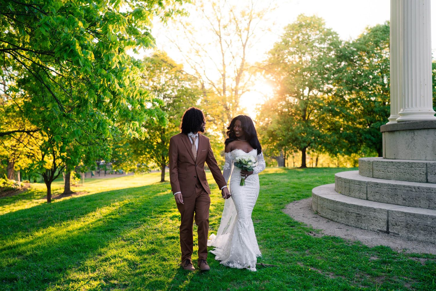 A Black couple in wedding attire walks hand-in-hand at sunset in a lush green park with golden light filtering through trees.