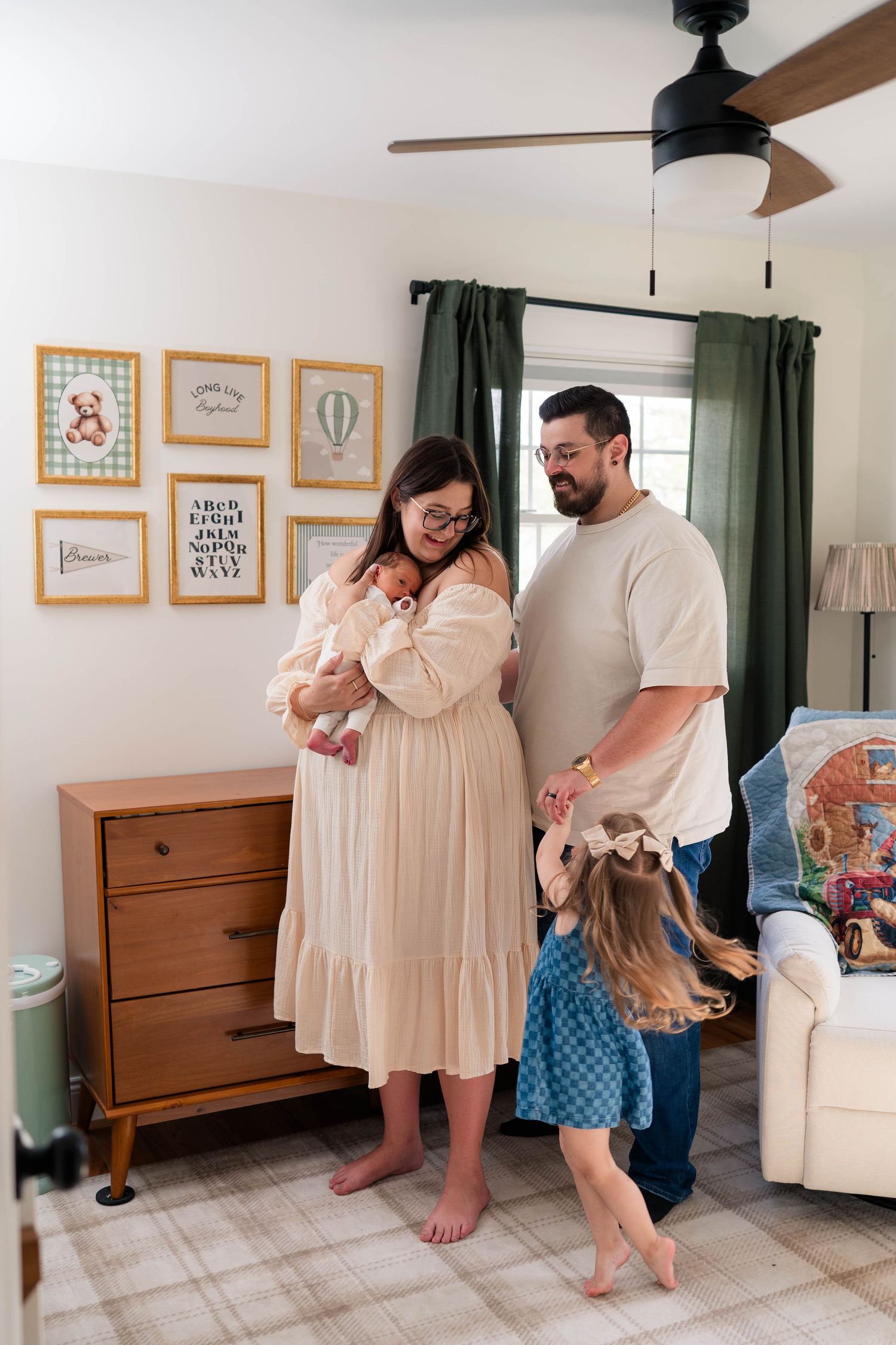 Young family with newborn and toddler daughter stands in a decorated nursery with gallery wall.