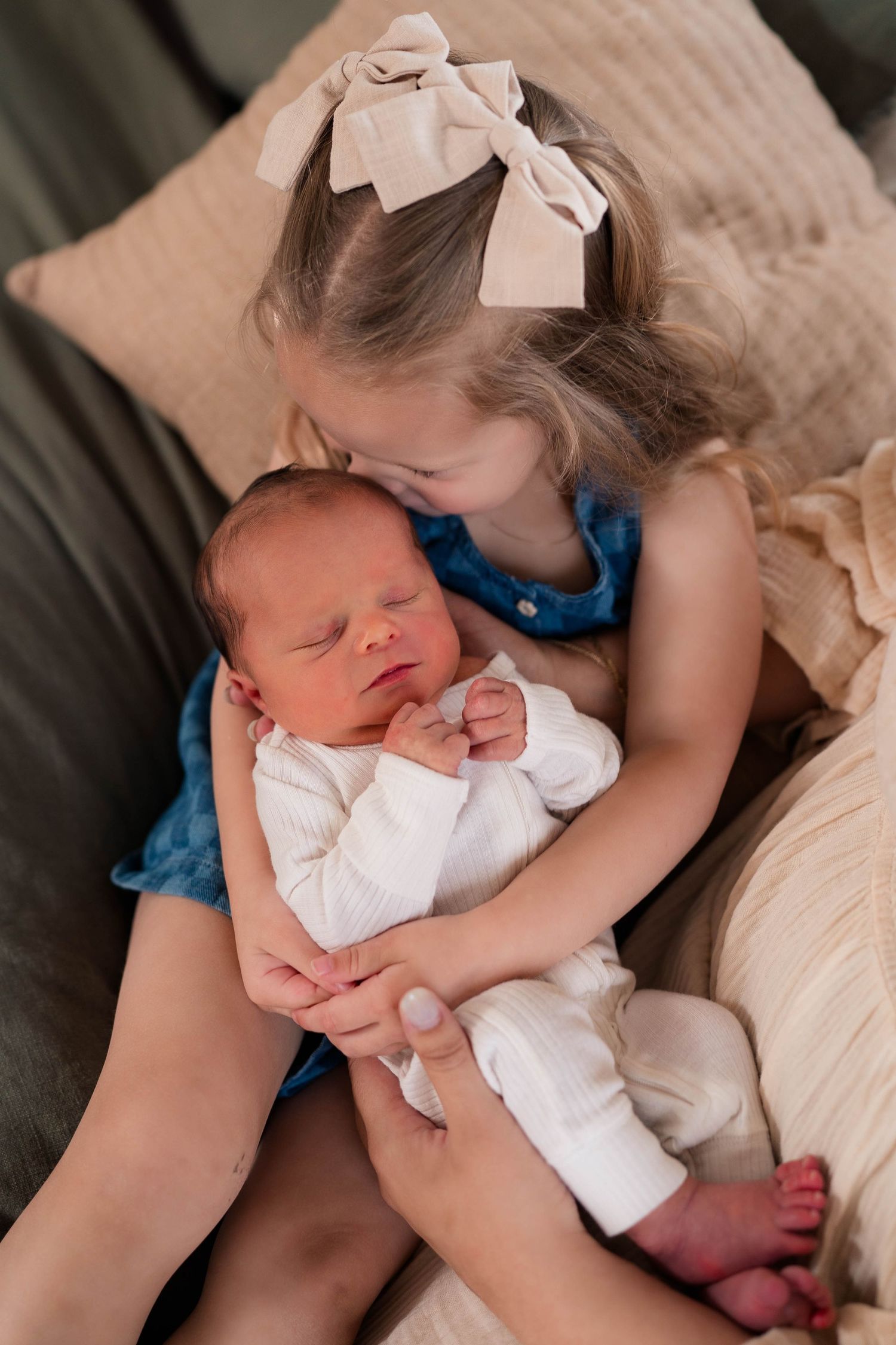 Young girl with bow kisses sleeping newborn baby sibling while holding them gently on a couch.