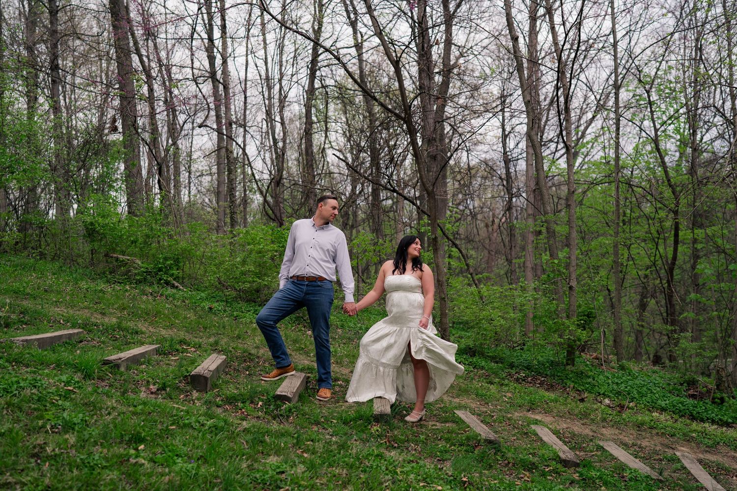 Couple holding hands walking down wooden steps through lush green forest.