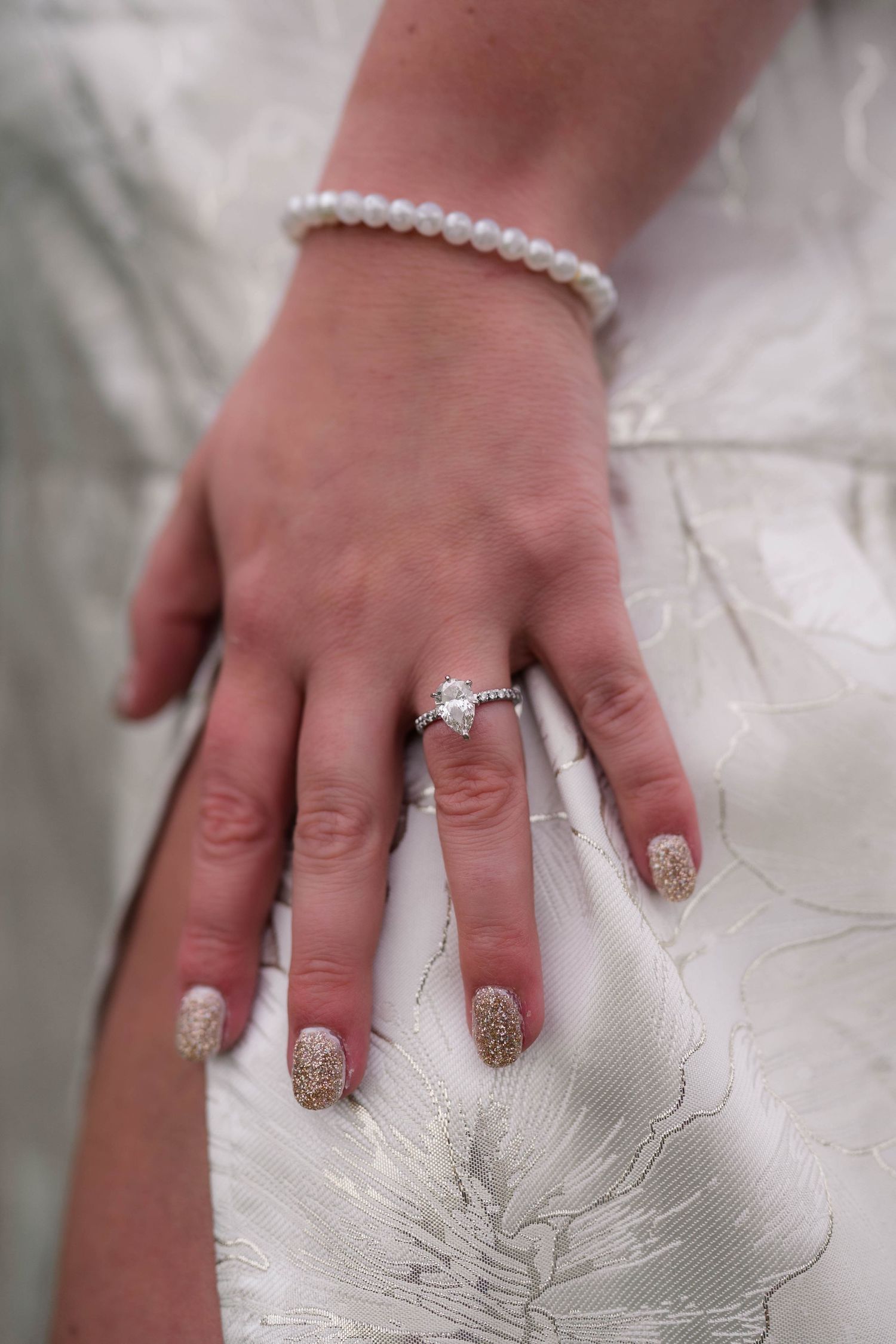 Close-up of bride's hand showing diamond engagement ring and pearl bracelet on white gown.