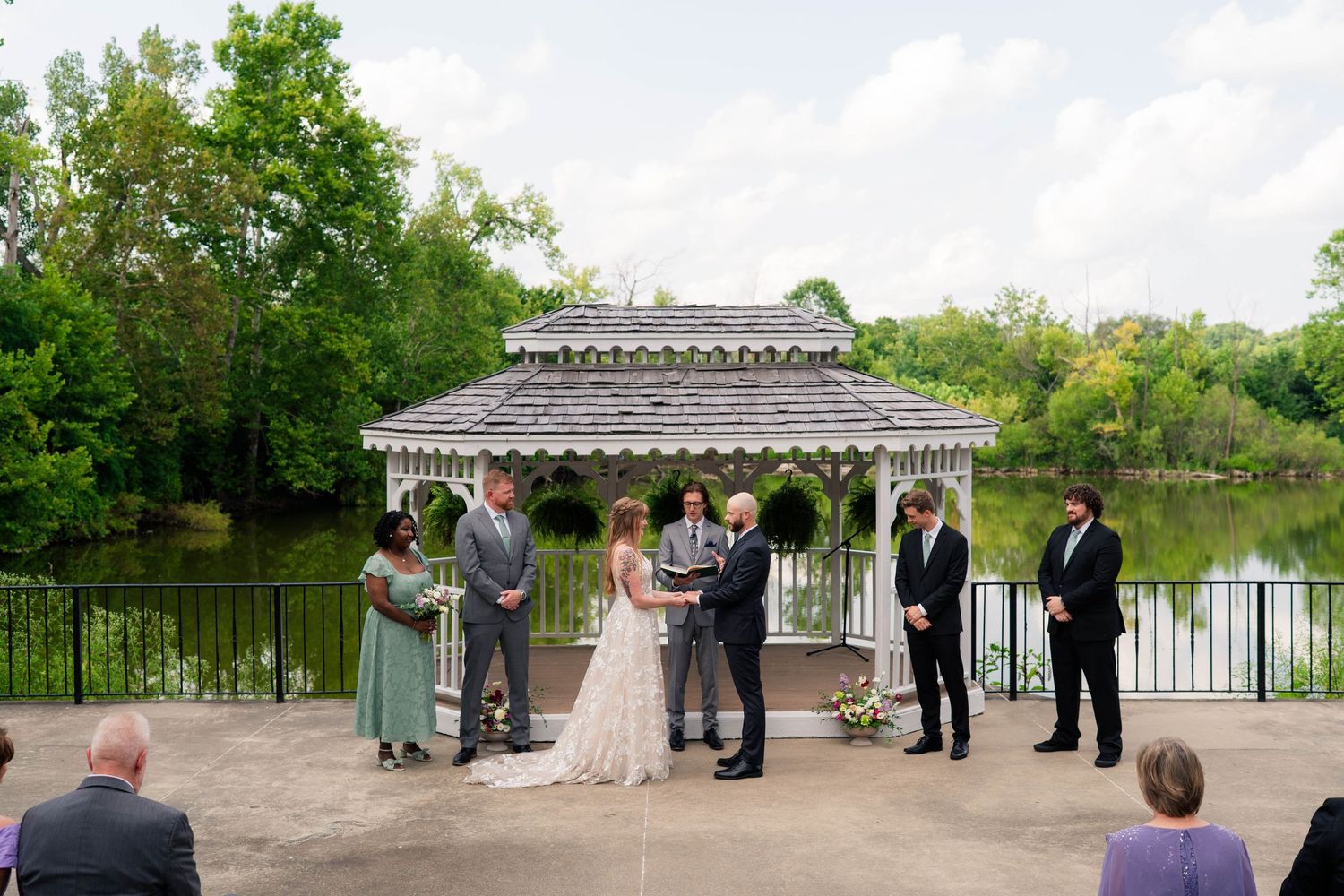 Outdoor wedding ceremony at gazebo by lake with bride, groom, and small wedding party.