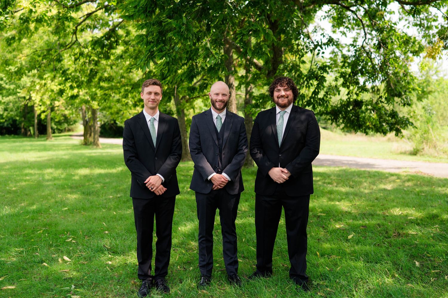 Three groomsmen in black suits with sage green ties stand together smiling in lush green park.