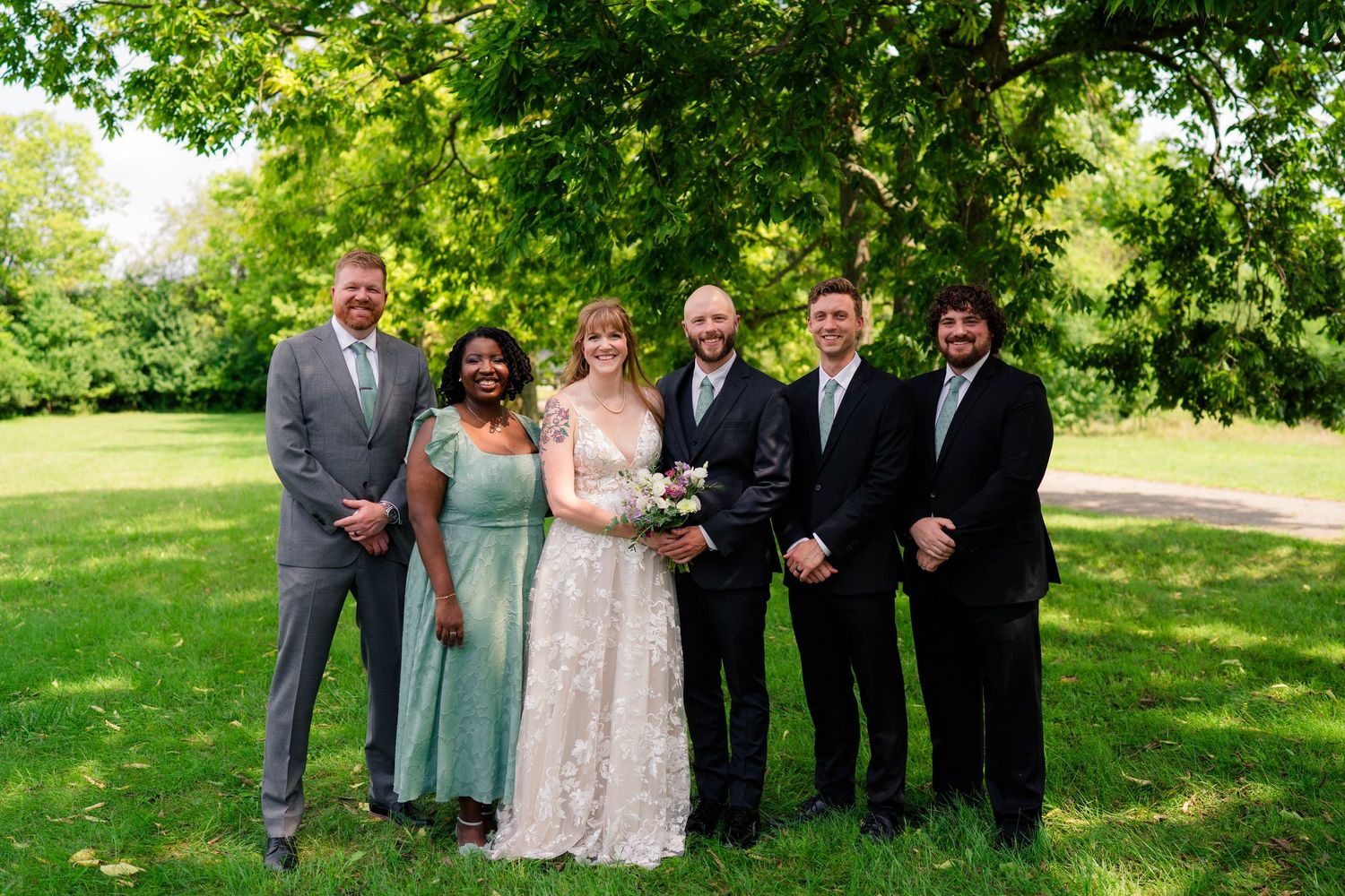 Wedding party group portrait with bride, groom, and four guests smiling under lush green trees.