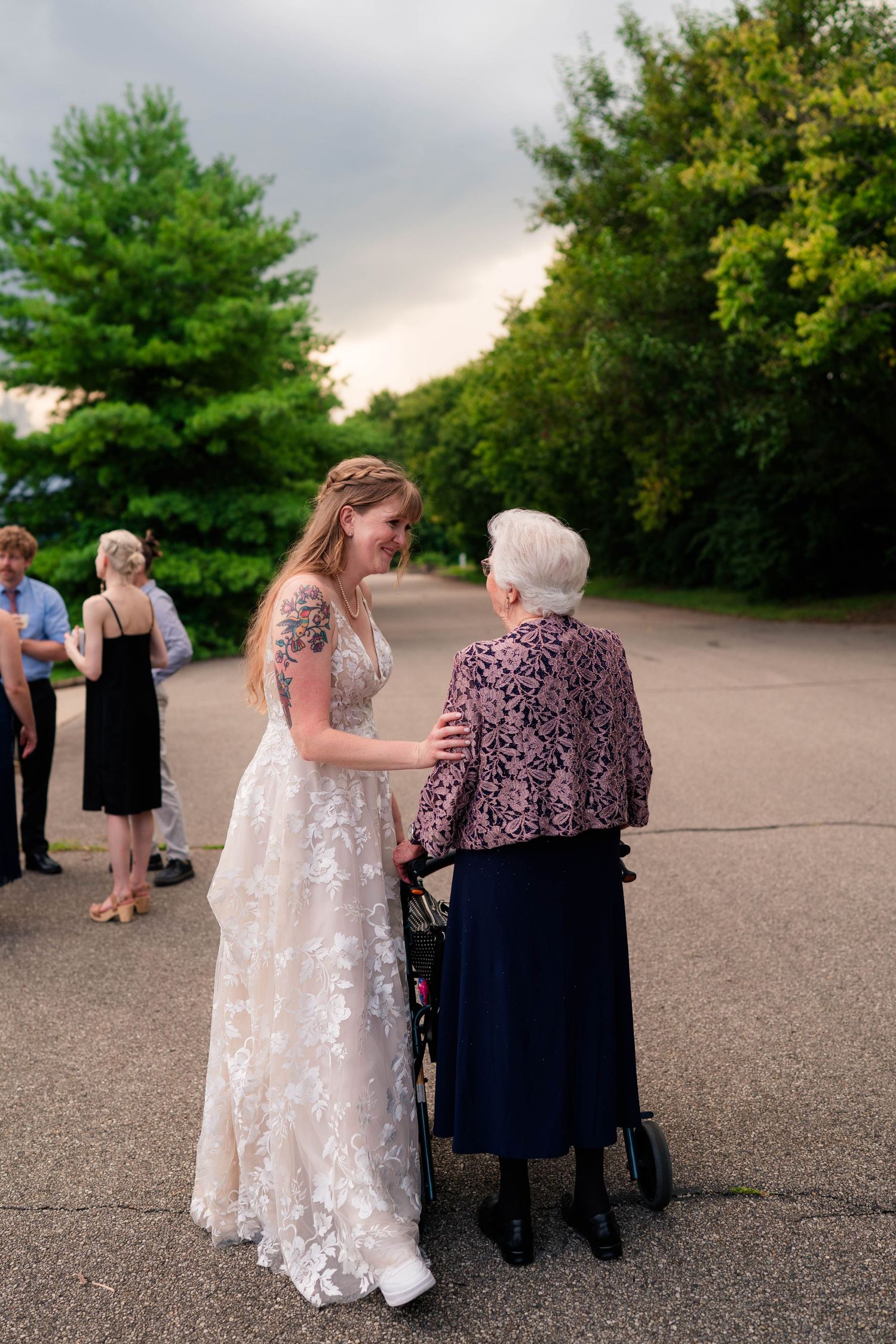 Bride in a floral lace gown tenderly interacts with an elderly woman using a walker at an outdoor wedding.