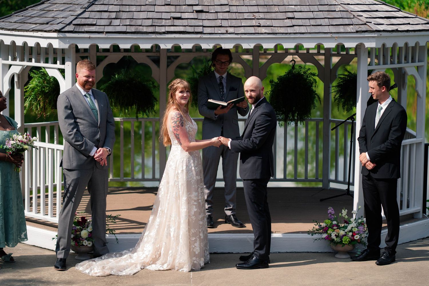 A bride and groom exchange vows at a white gazebo altar flanked by their wedding party outdoors.