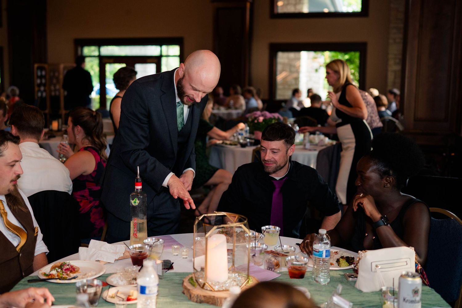 Bald groom in suit greets seated guests at wedding reception dinner.