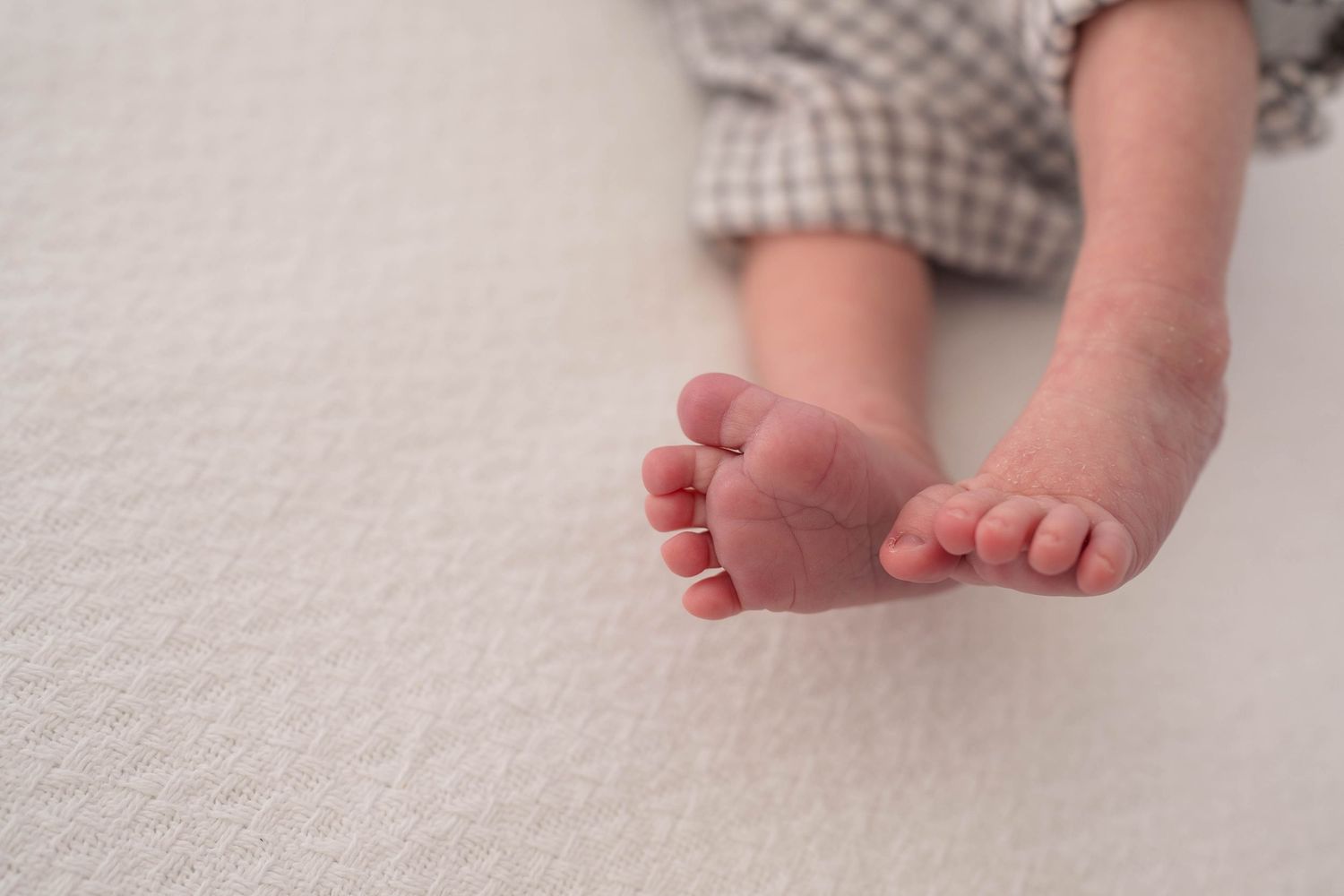 Close-up of tiny newborn baby feet resting on a soft white blanket, wearing plaid pants.