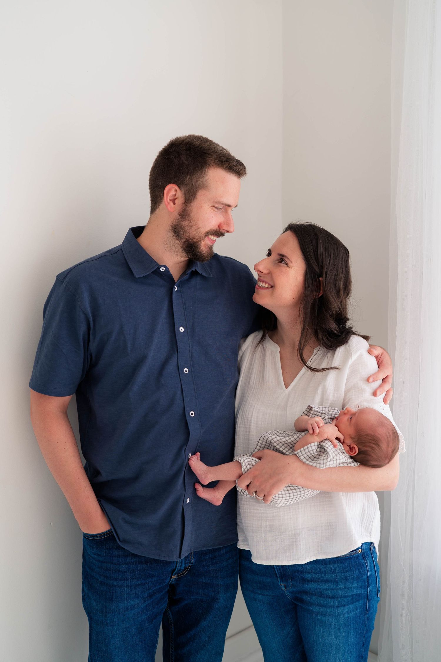 Smiling couple in casual attire gazing at each other while holding their sleeping newborn baby against a white wall.