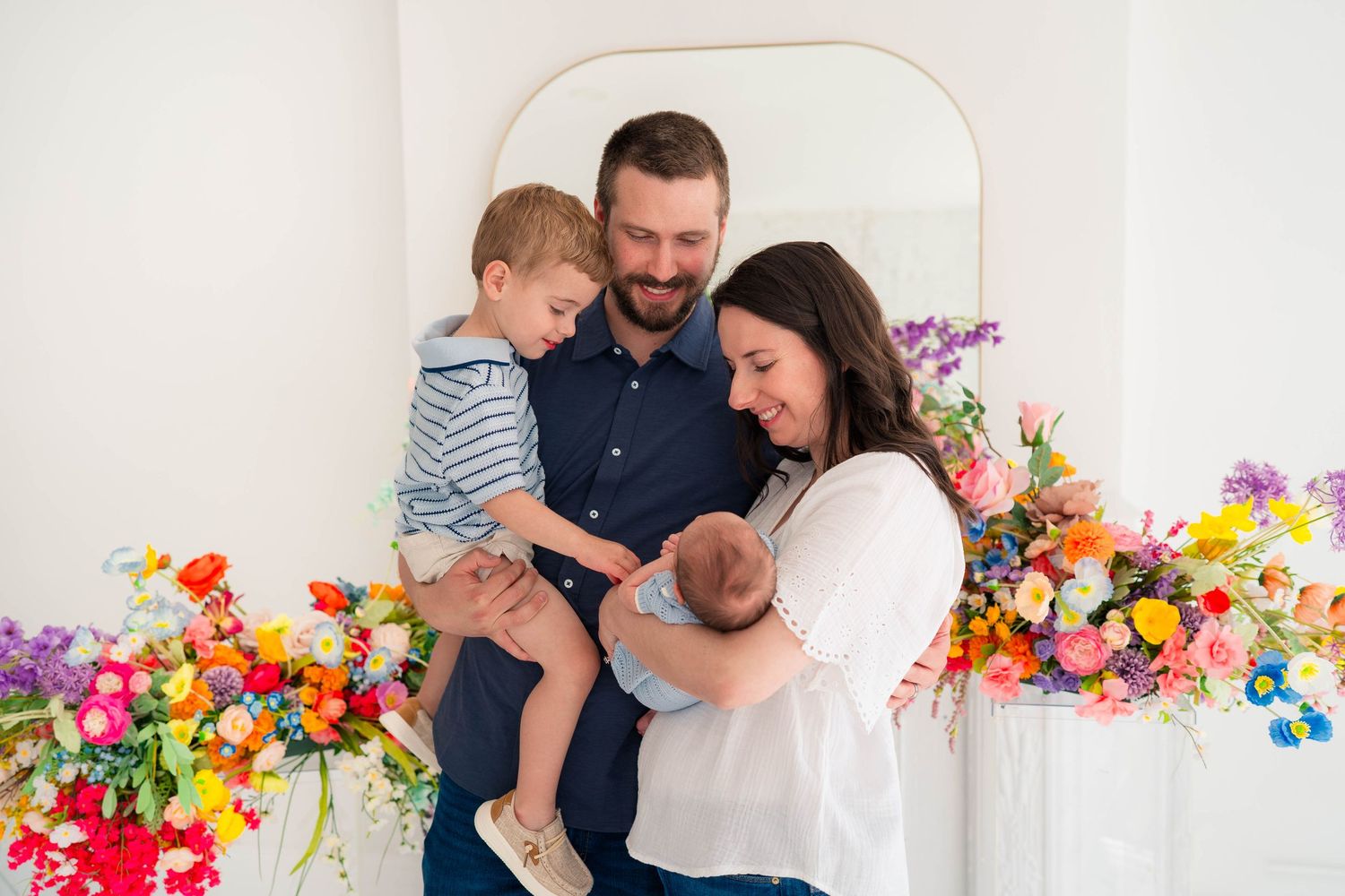 Family of four with toddler and newborn smiling together amid vibrant flowers.