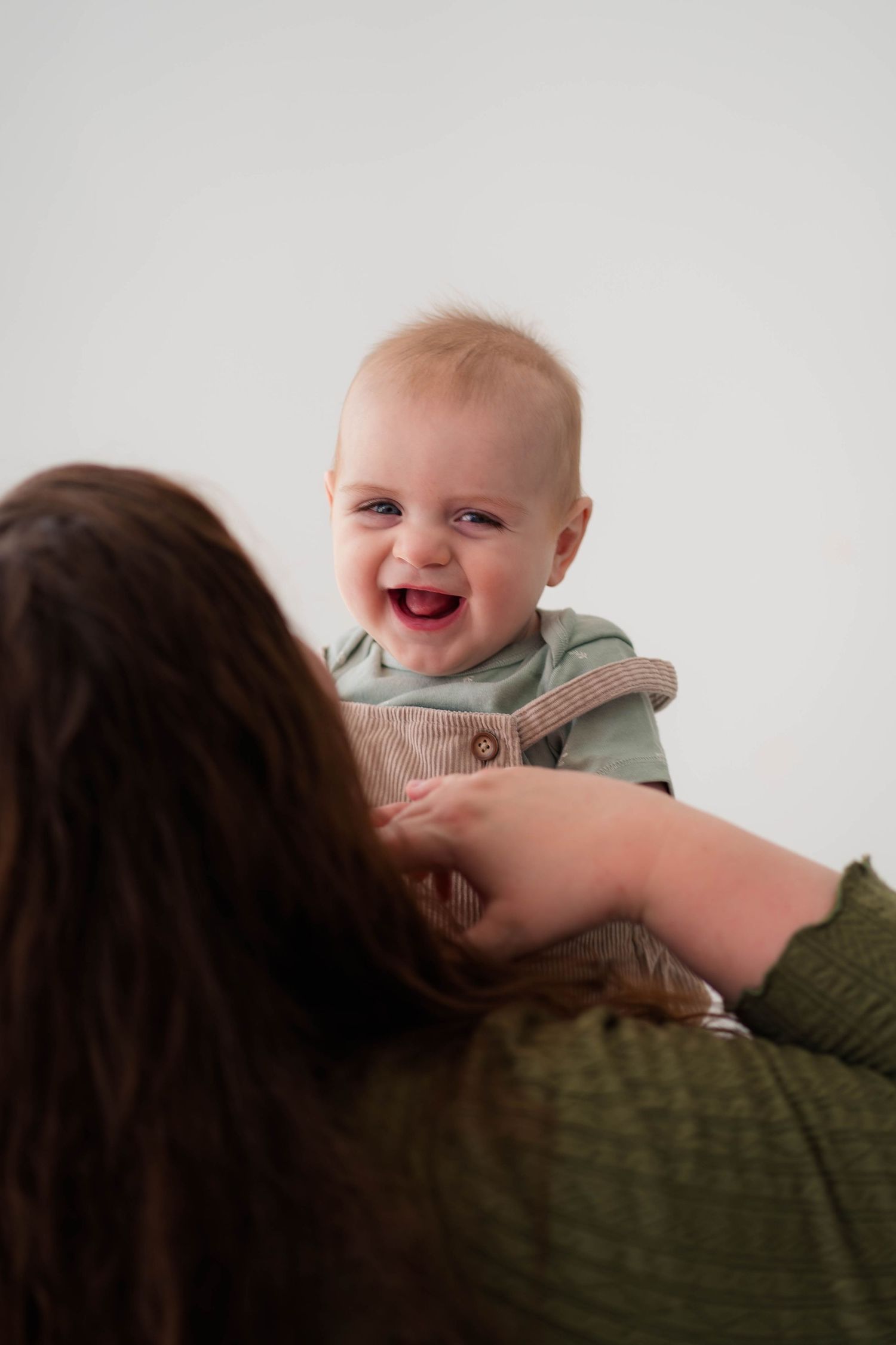 Joyful baby laughing while being held up by mother in green sweater, studio photo.