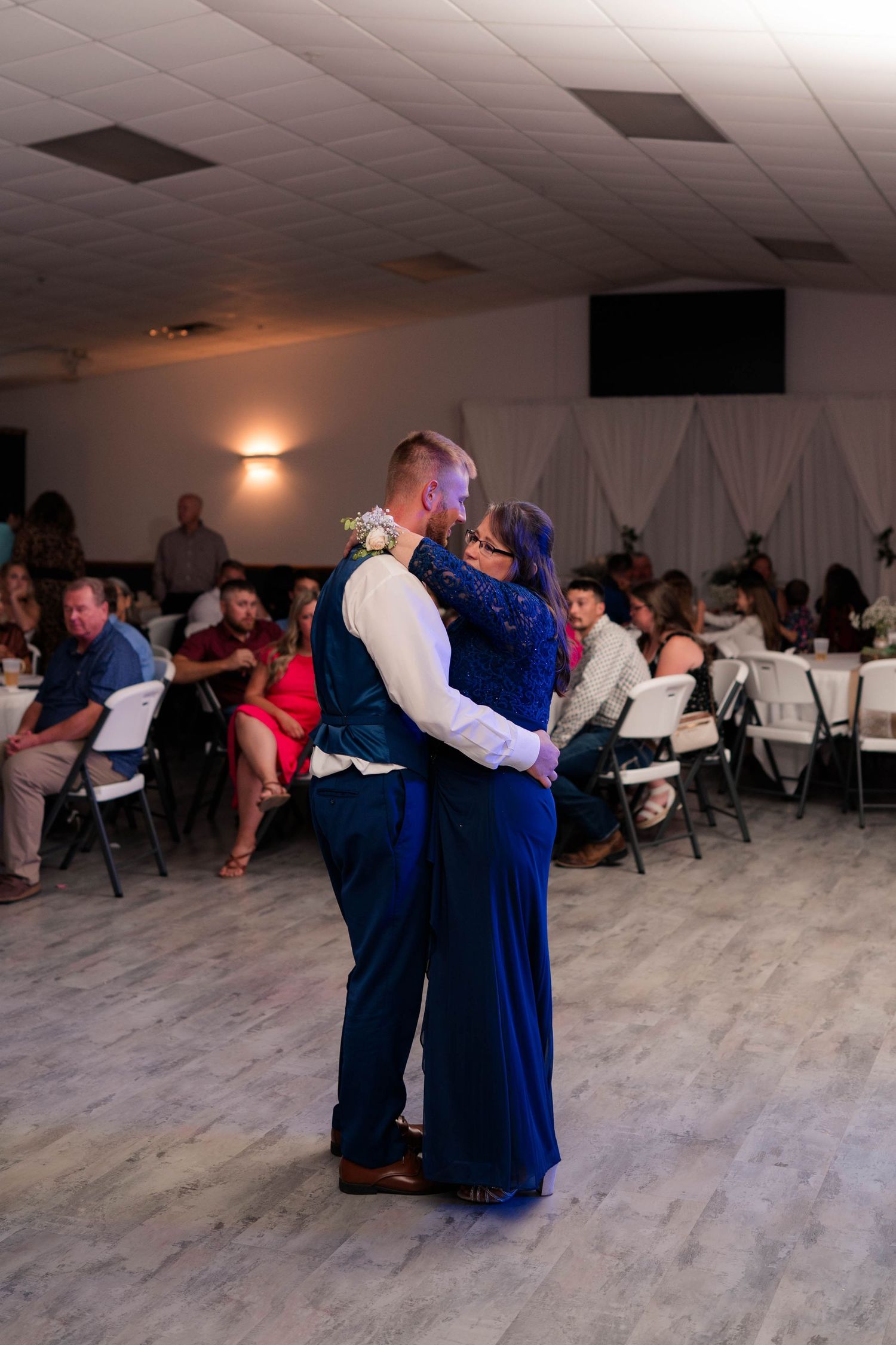 Groom and mother share a tender dance on the floor at a wedding reception with guests watching.