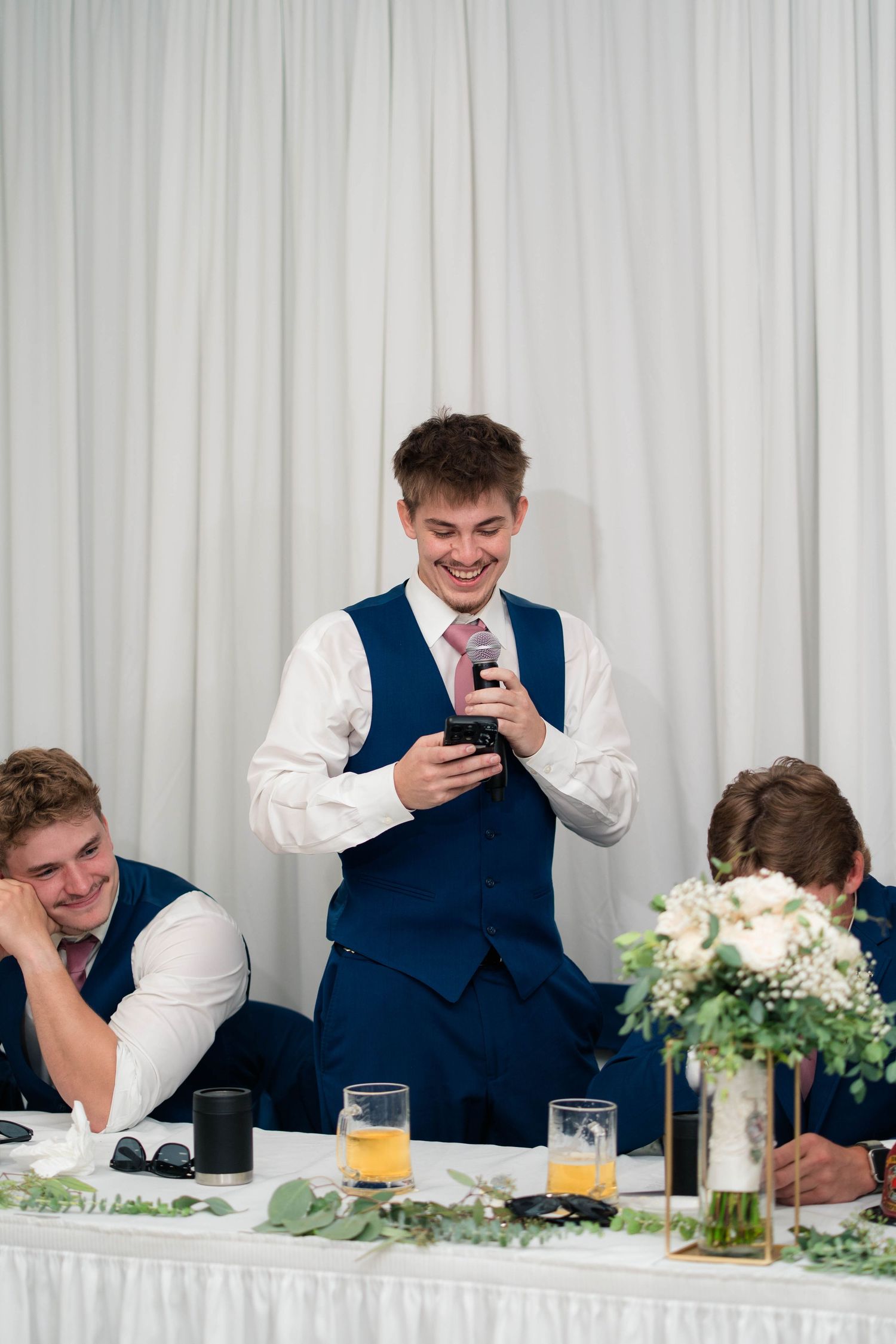 Smiling groomsman in navy vest delivers wedding speech holding microphone at elegant reception.