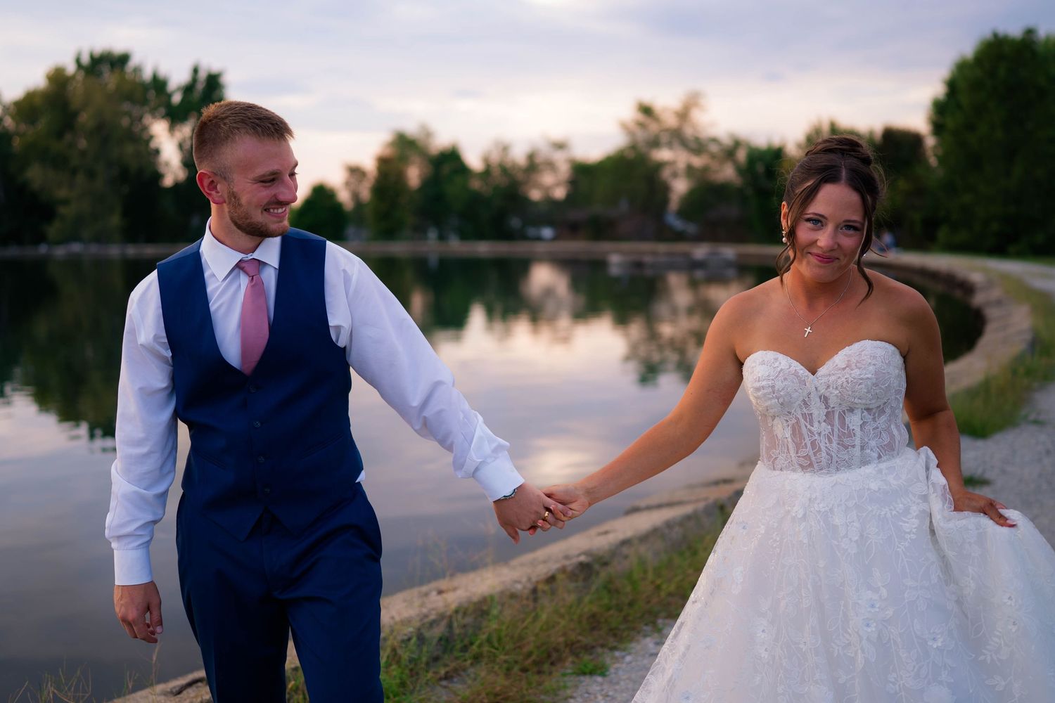 Bride and groom hold hands by a lakeside at sunset, she in a lace gown, he in a navy suit with pink tie.